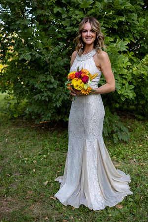 Bride in lace dress holds colorful sunflower bouquet against green garden backdrop.
