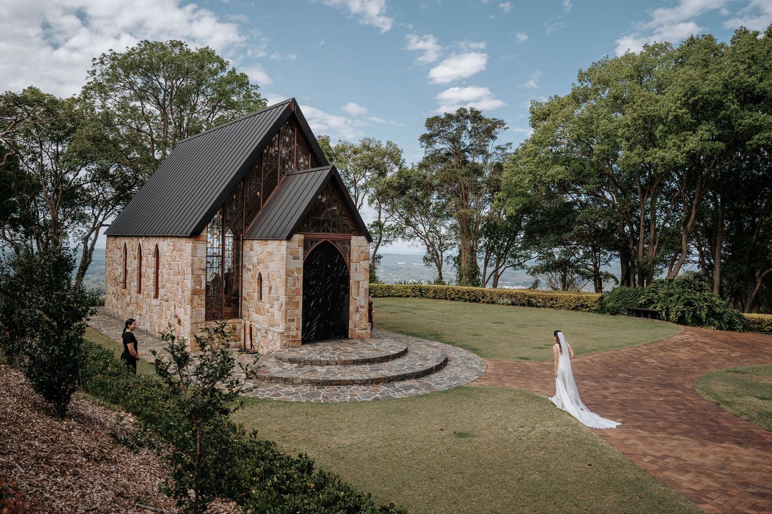 A rustic stone chapel with a steep roof sits on a hilltop overlooking trees, with a figure in white standing nearby.