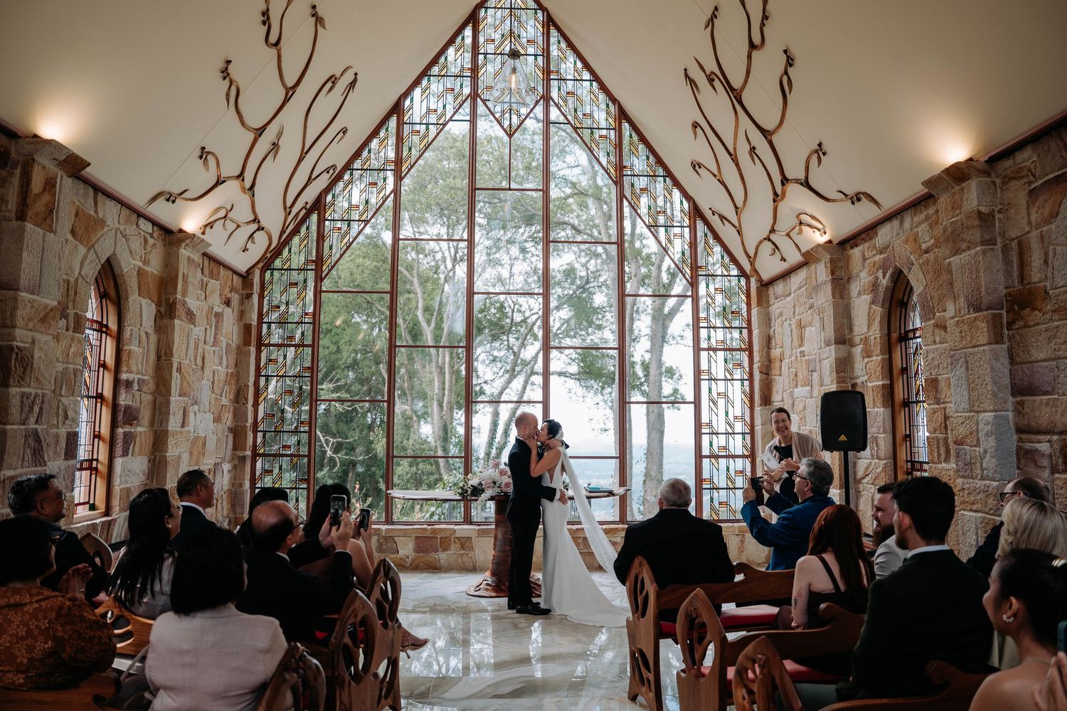 Wedding ceremony in a rustic stone chapel with large windows and natural light streaming through.