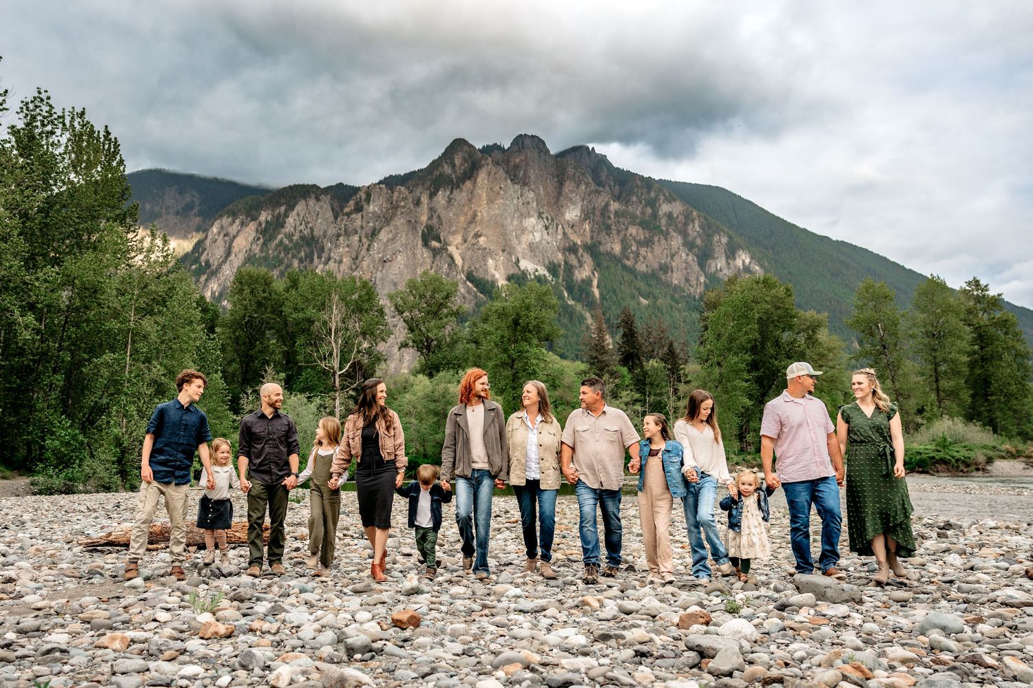 Large group gathered on rocky riverbank with mountain backdrop.