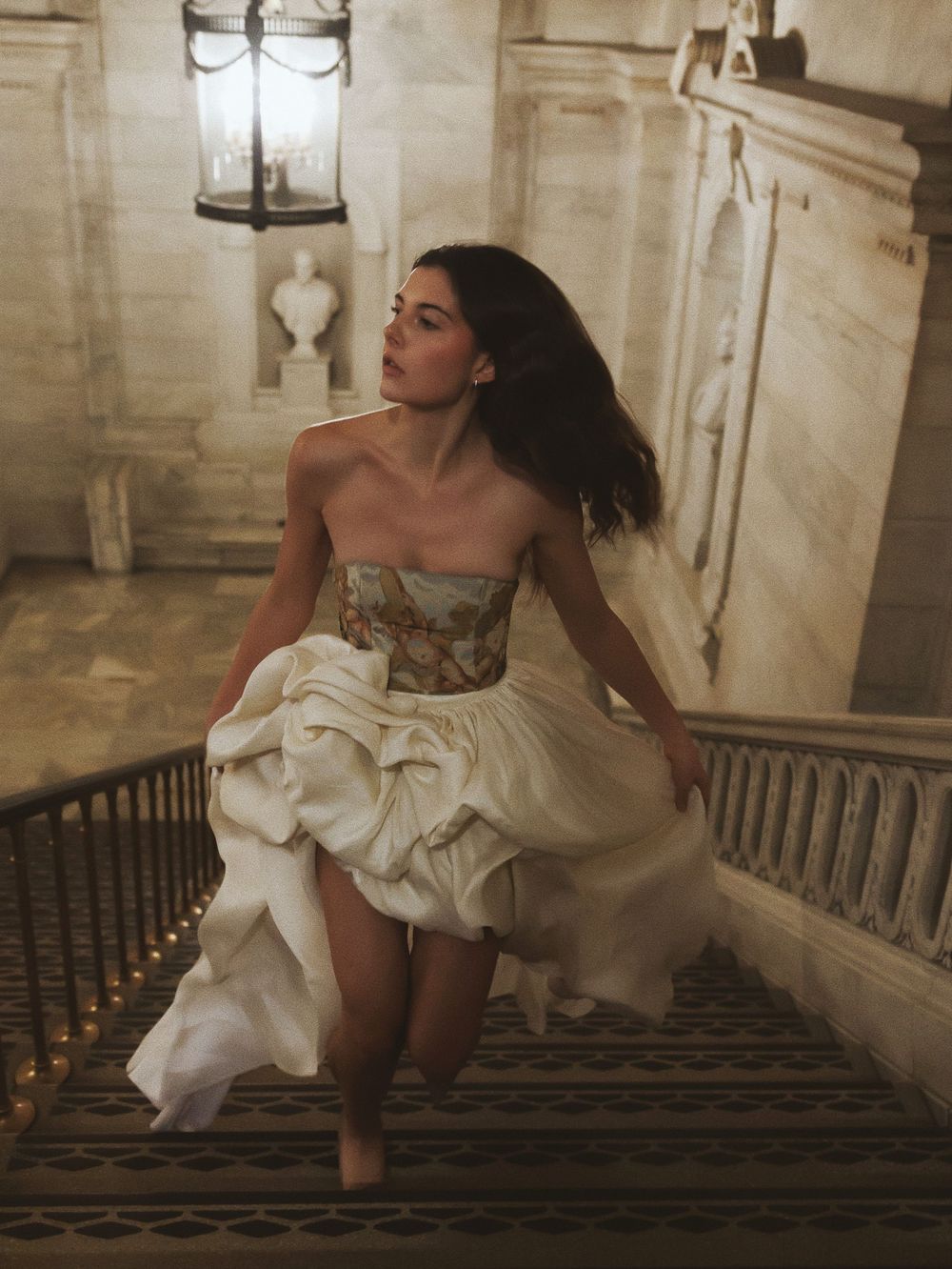 An artistic fashion photoshoot on grand marble stairs showcases a flowing white gown that moves elegantly with the model.