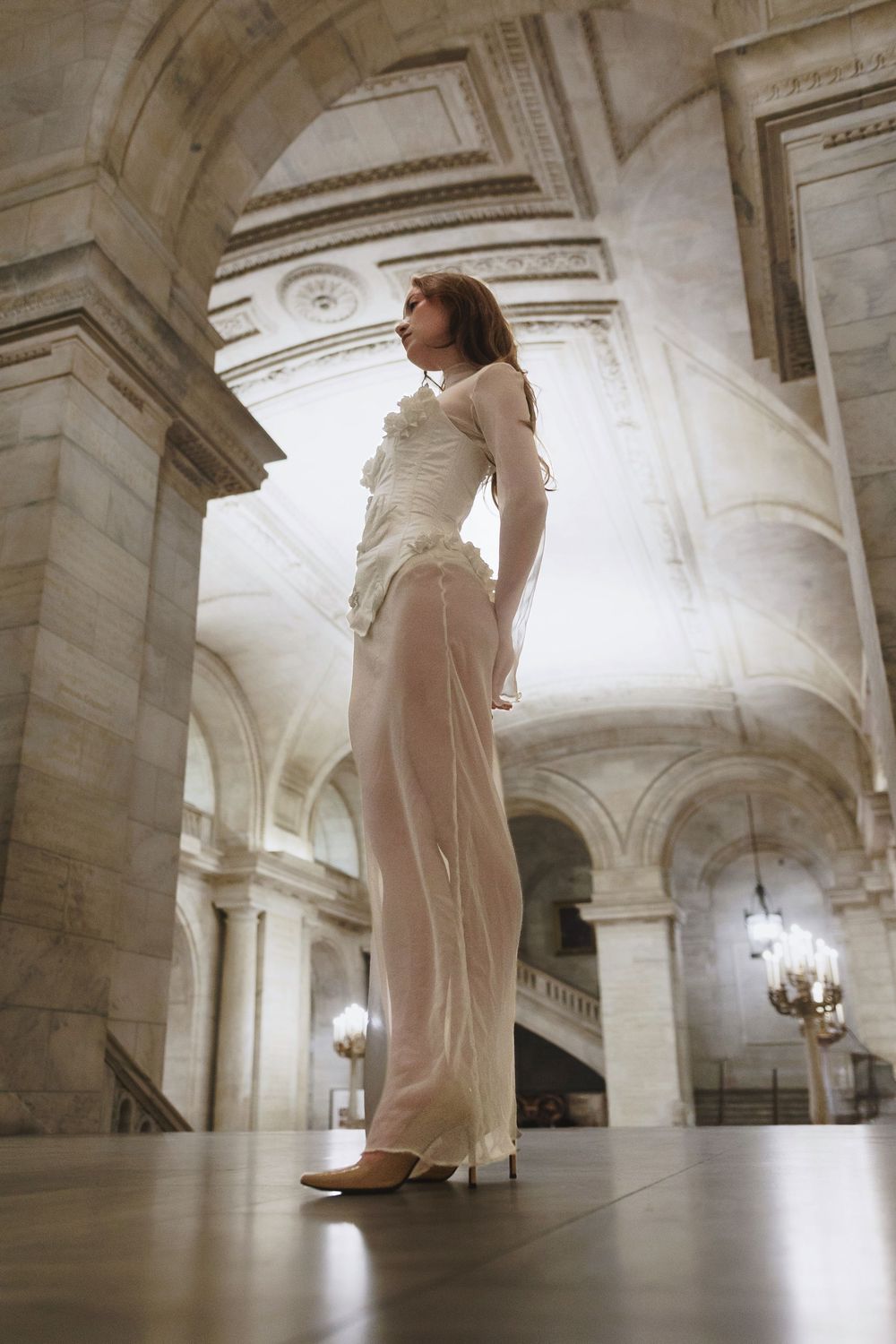 A figure in an elegant white outfit stands in a grand architectural space with arched ceilings and marble columns.