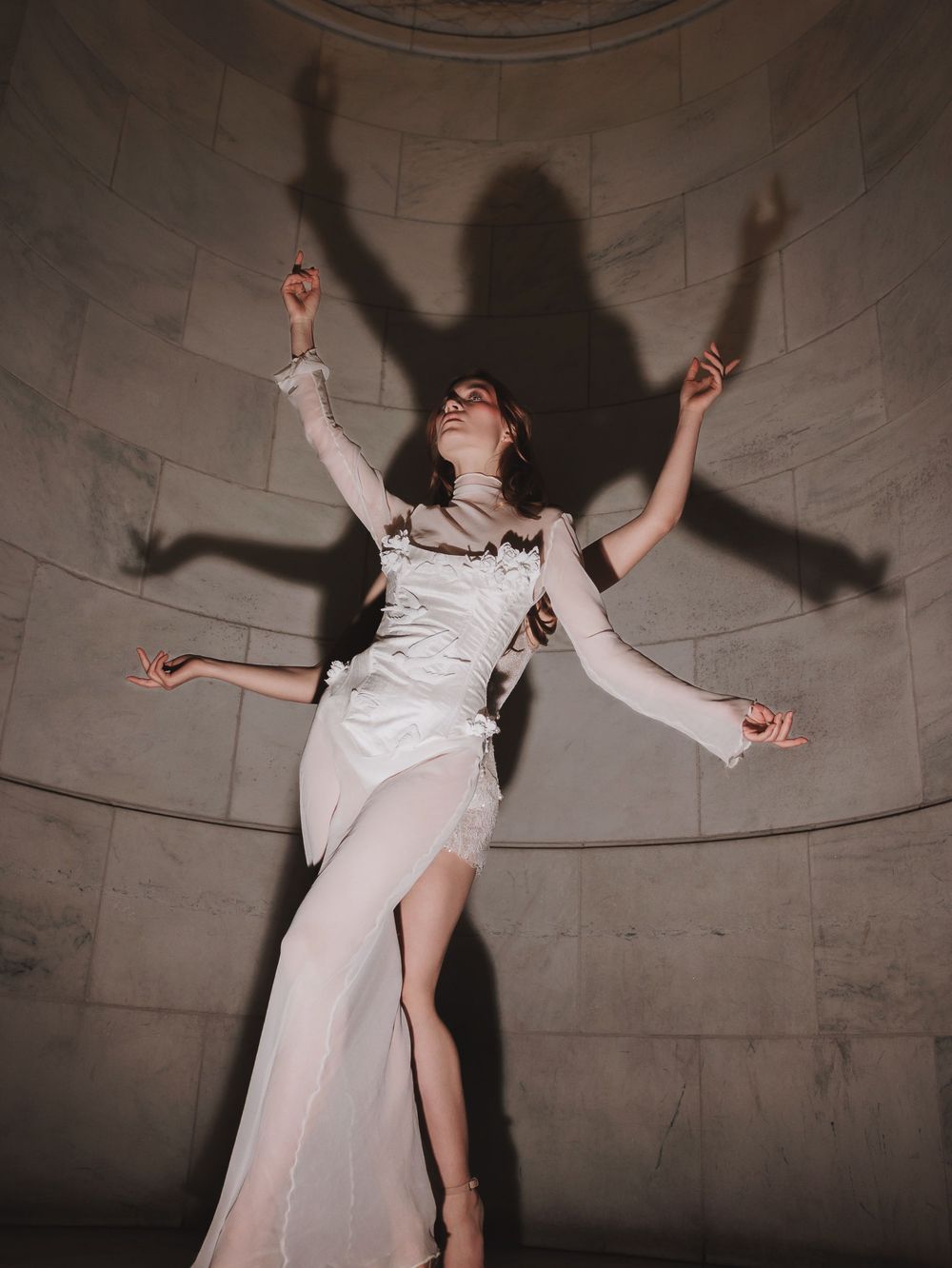 A figure in a flowing white dress dances expressively against a grey stone wall, casting dramatic shadows in multiple poses.