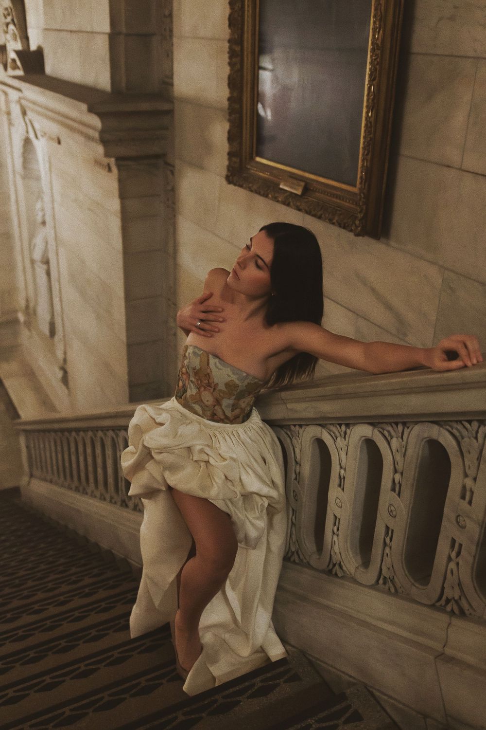 A graceful figure in a light-colored dress reclines dramatically against an ornate architectural balustrade.