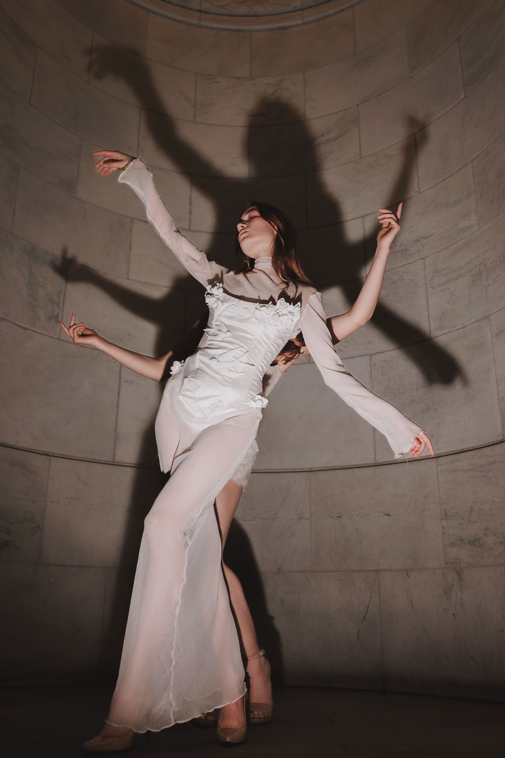 A person in white flowing clothing dances and leaps expressively against a textured gray wall, creating dramatic shadows.
