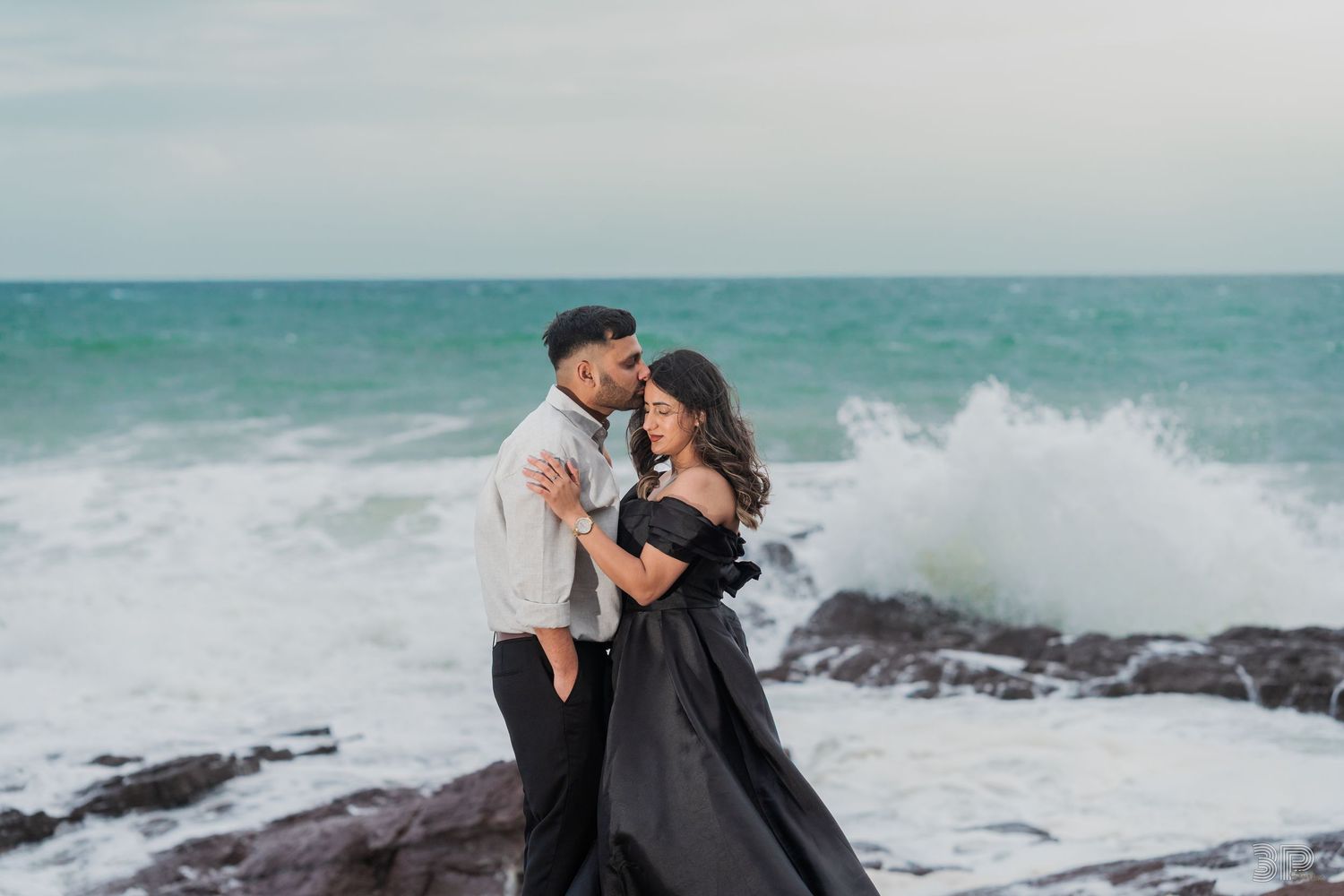 A couple shares tender moments along a rocky beach coastline with crashing waves in the background.
