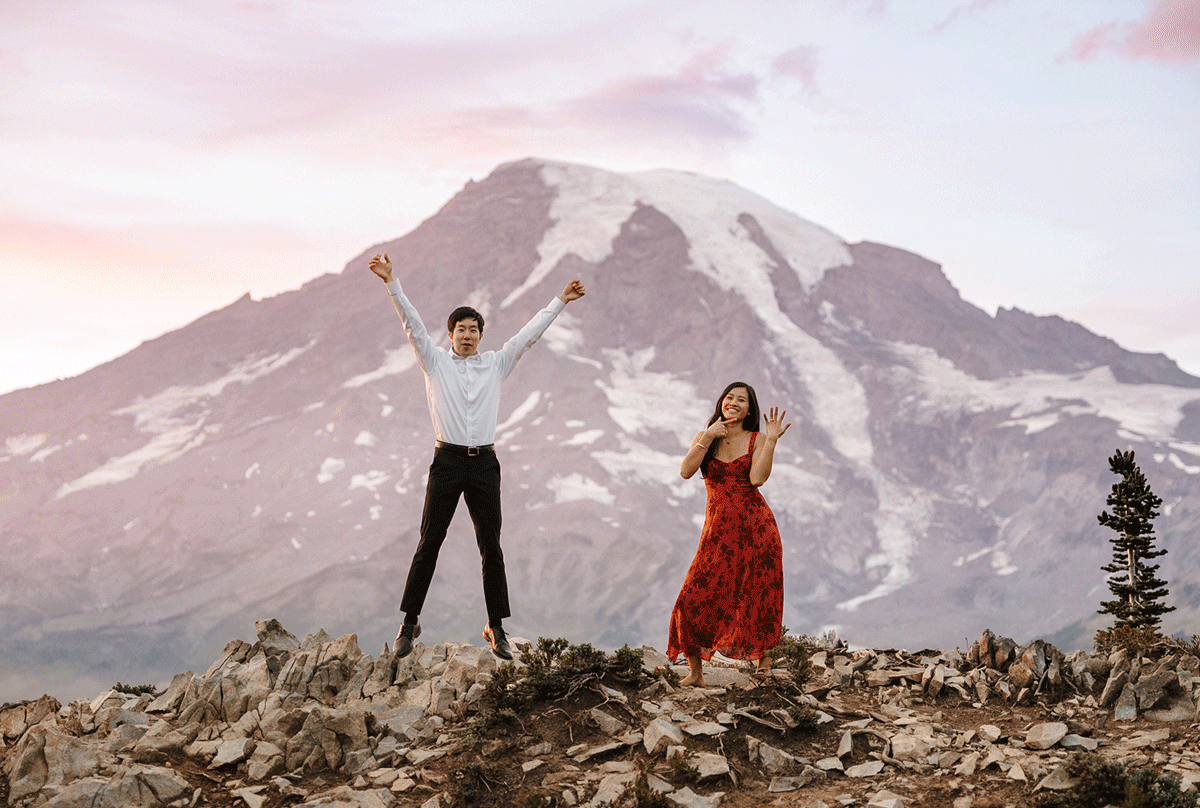 Two figures stand triumphantly with arms raised against a snow-capped mountain backdrop at sunset.