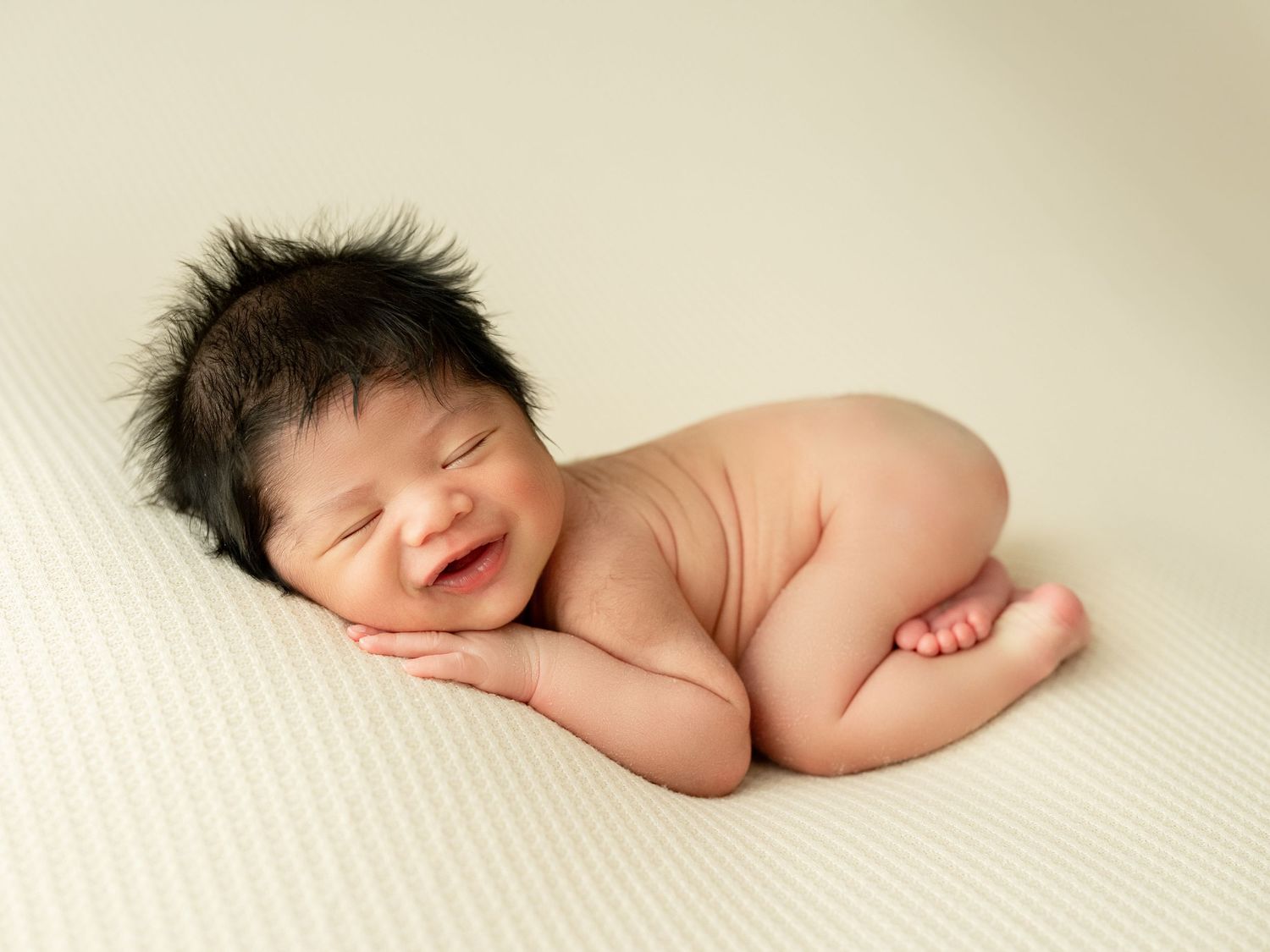 A sleeping newborn baby rests peacefully on a soft cream-colored blanket in a professional photography setting.