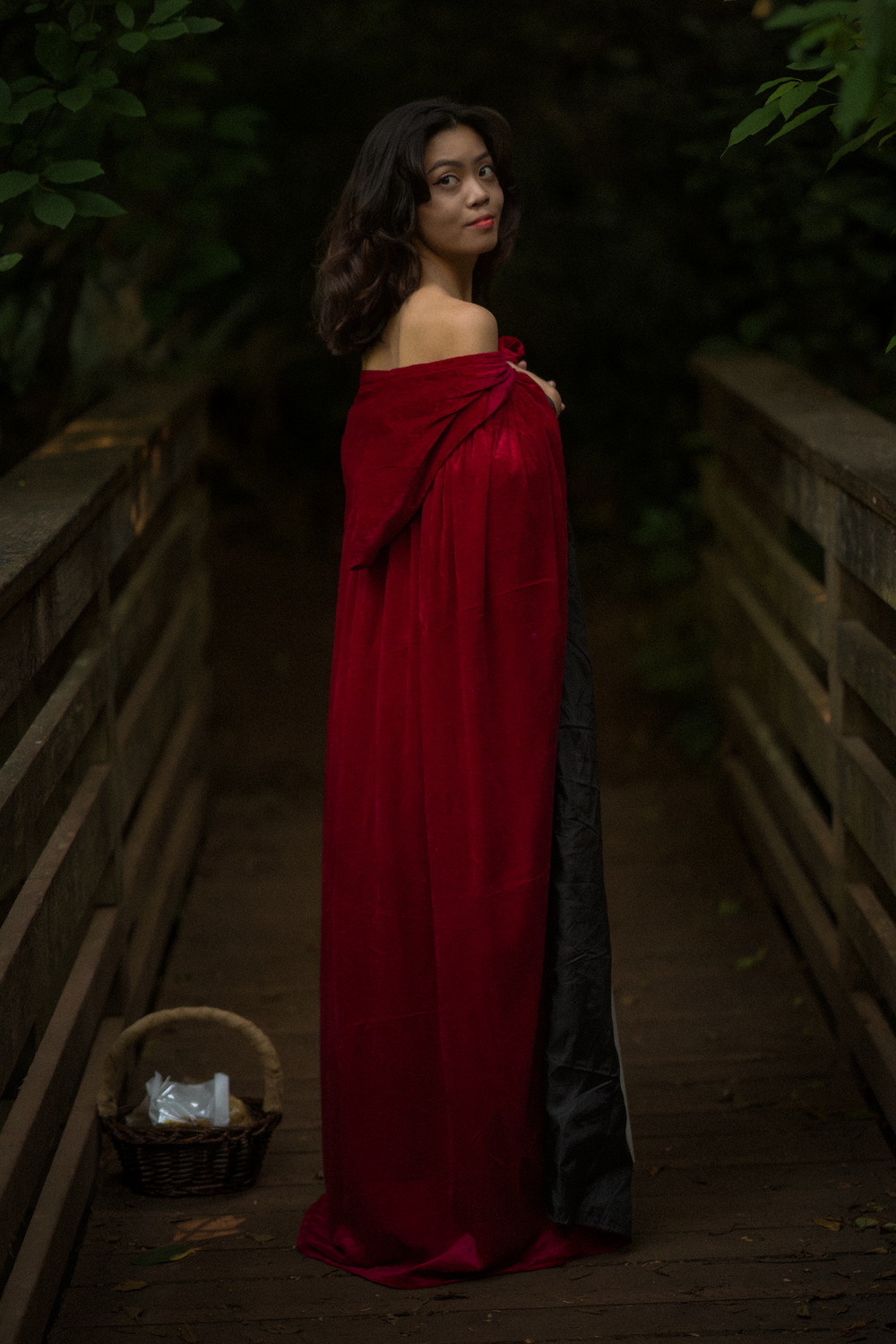 A figure in a red cloak and white dress poses dramatically on a woodland path in moody lighting.