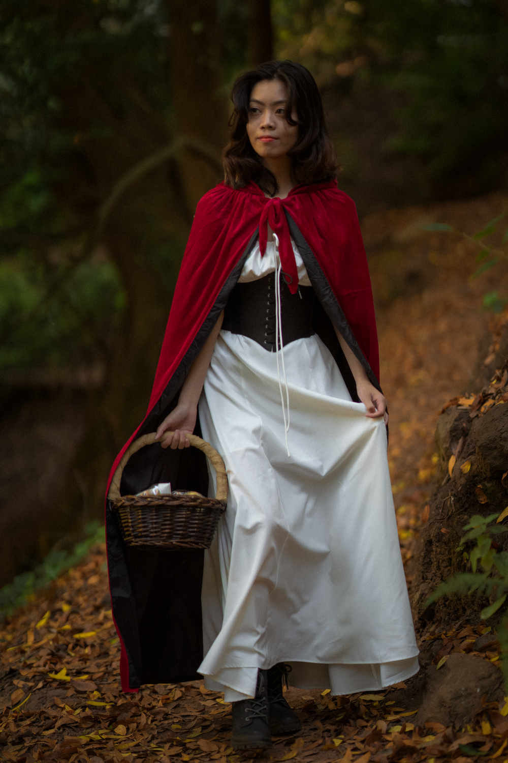 A person wearing a red cape and white dress sits on forest ground holding a woven basket.