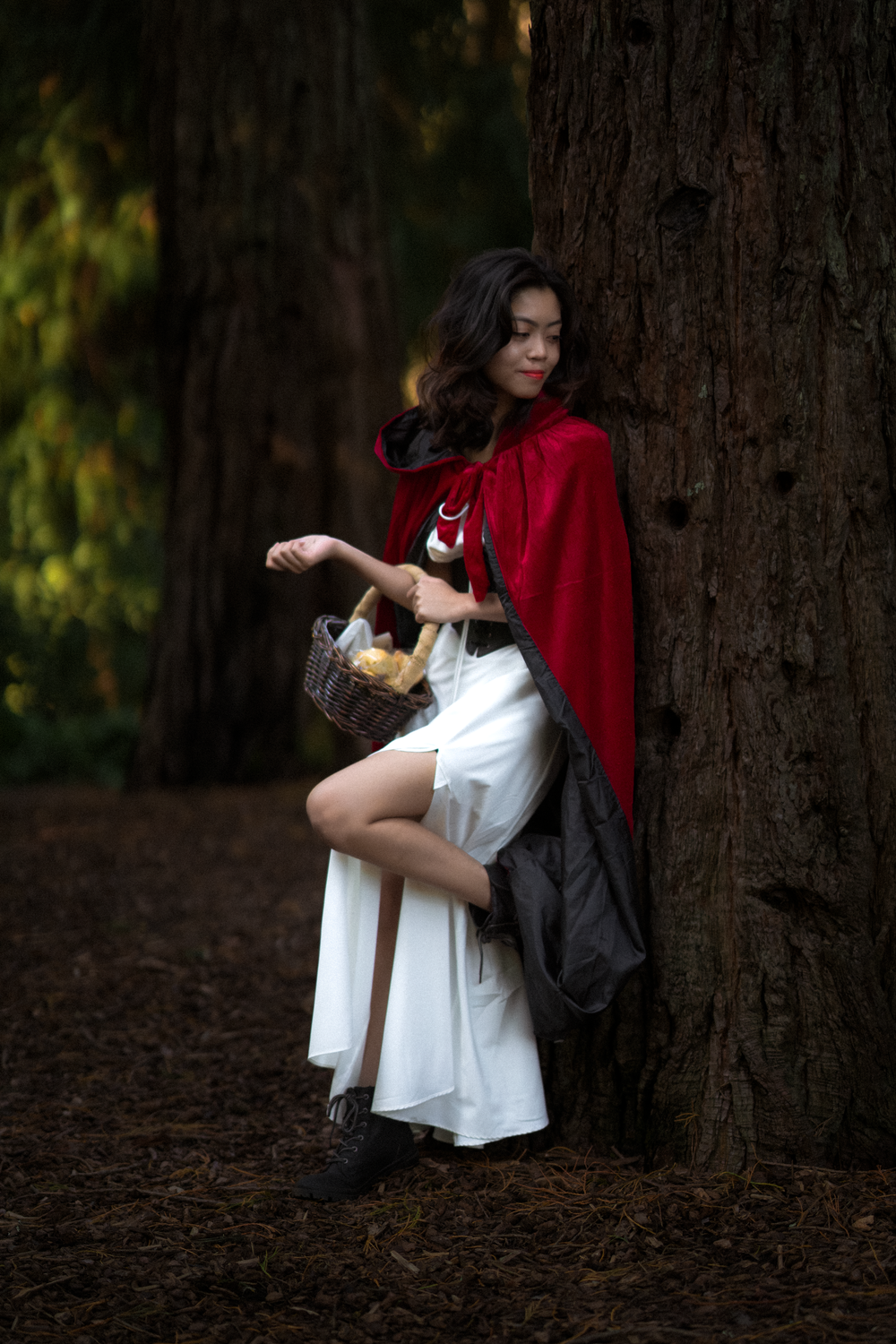 A person in a red cape and white dress poses near tree trunks in a dark forest setting.