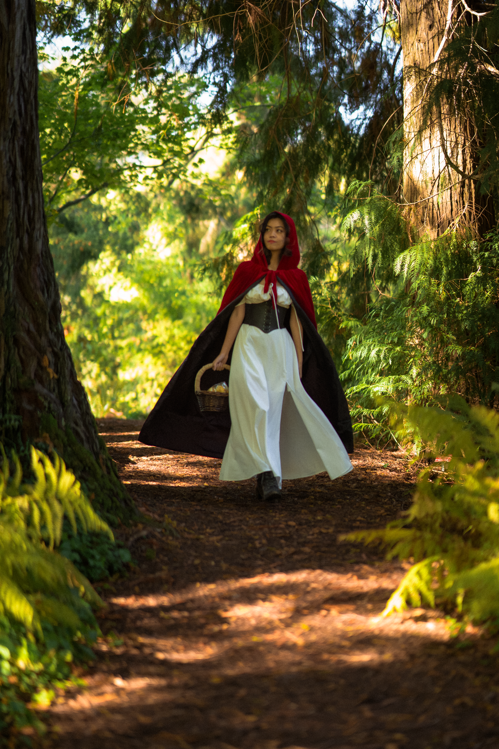 A person in a red cape walks along a sunlit forest path surrounded by tall trees.