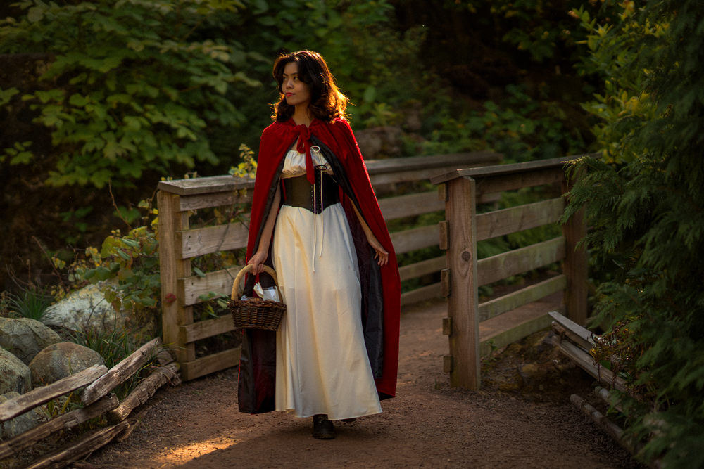 A figure in a red cape and white dress stands on a wooden bridge in a forest setting.