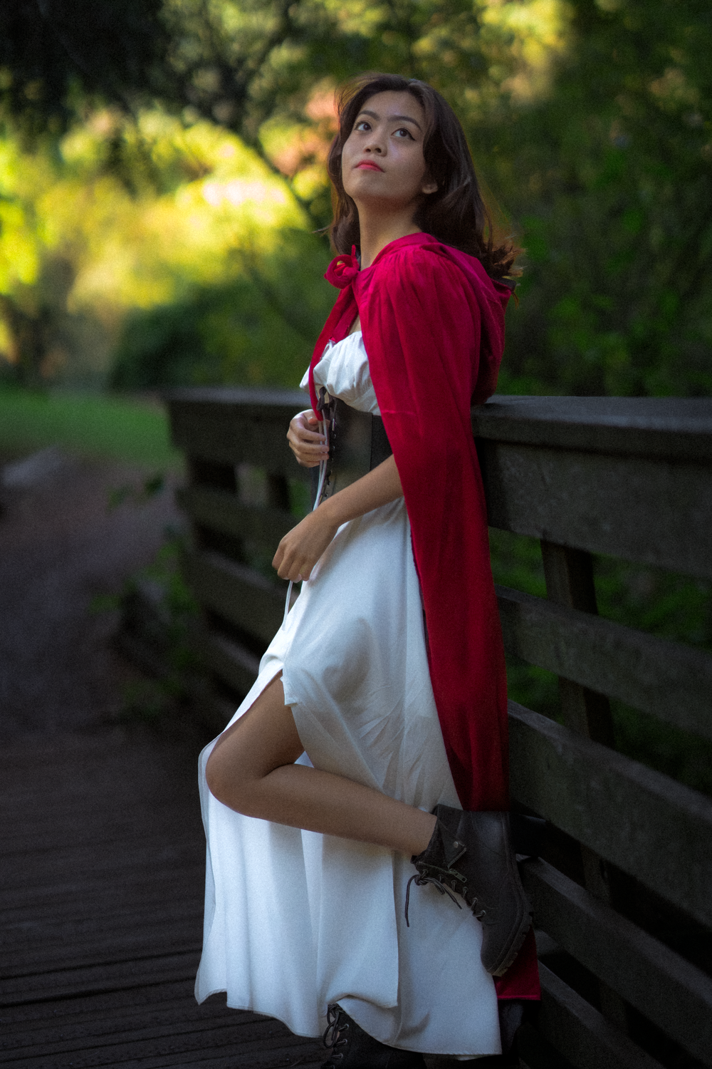 A person in a flowing white dress and red cape poses on a wooden bridge in dappled sunlight.