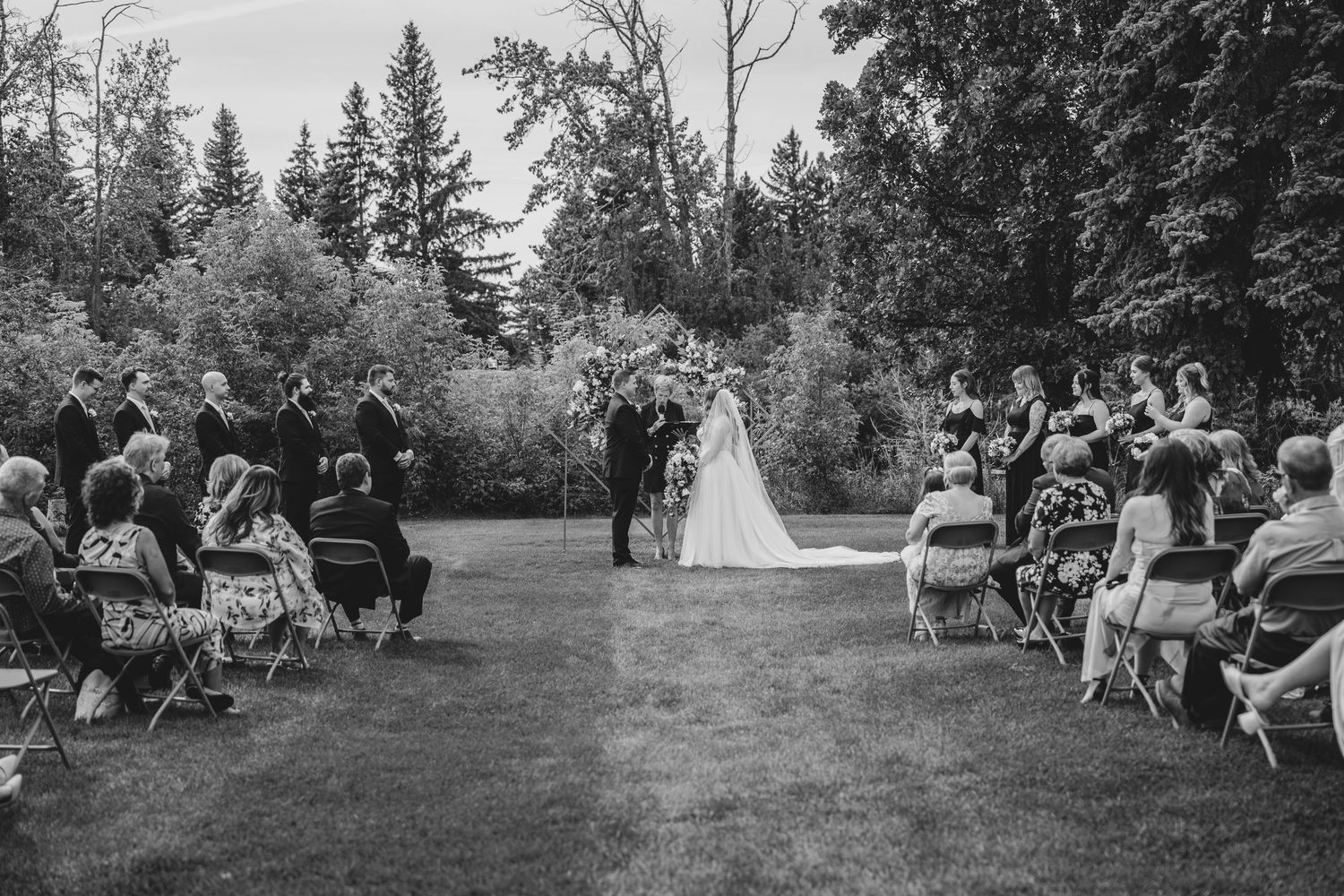 Black and white panoramic photo showing an elegant outdoor wedding ceremony surrounded by tall trees and seated guests.