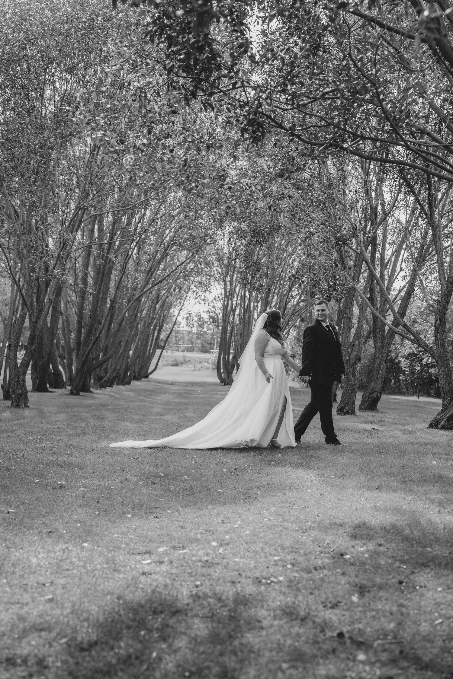 Wedding couple walks through a scenic tree-lined path in black and white photography.