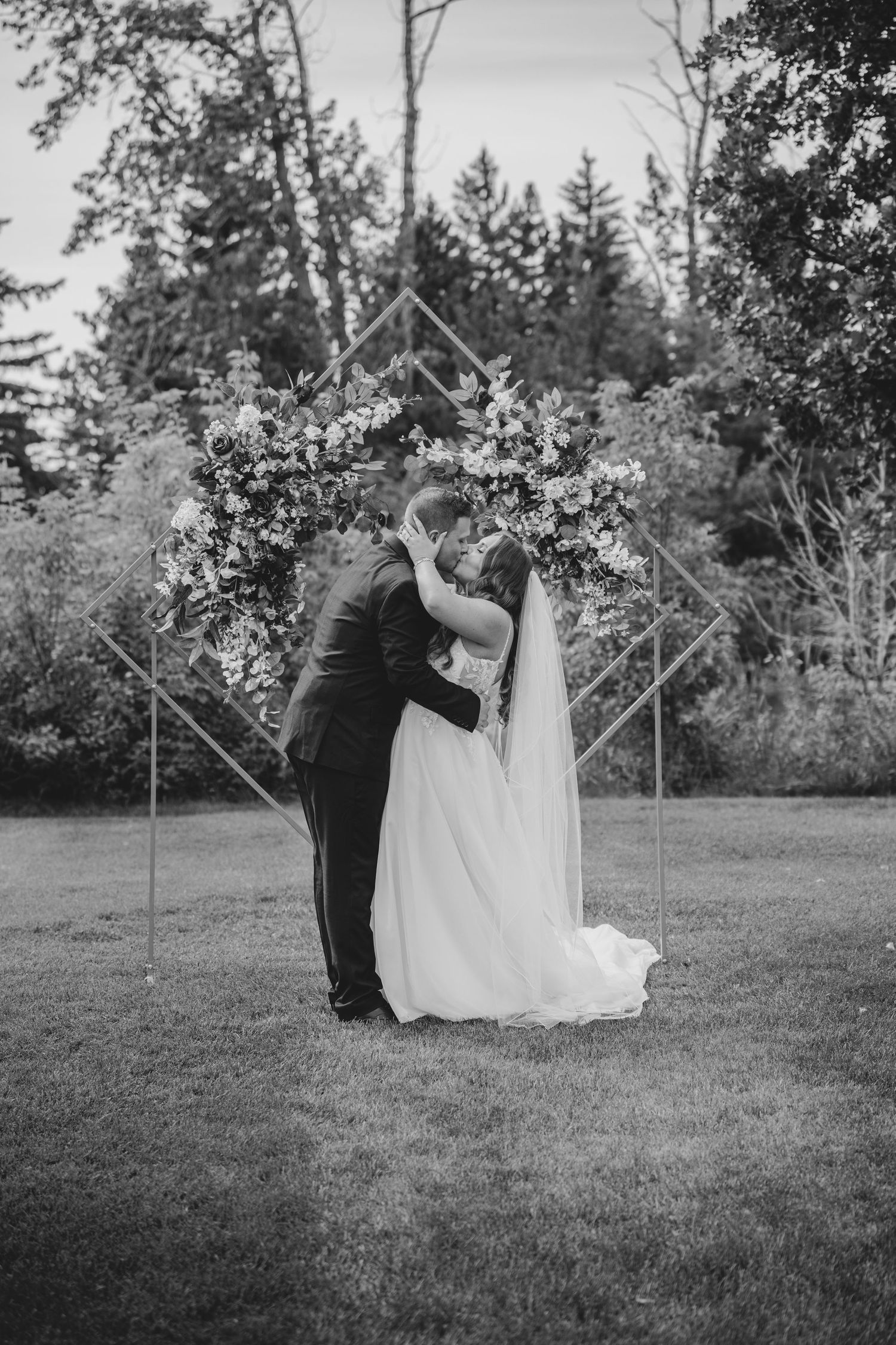 Couple in wedding attire dances and twirls on a grassy lawn surrounded by trees.