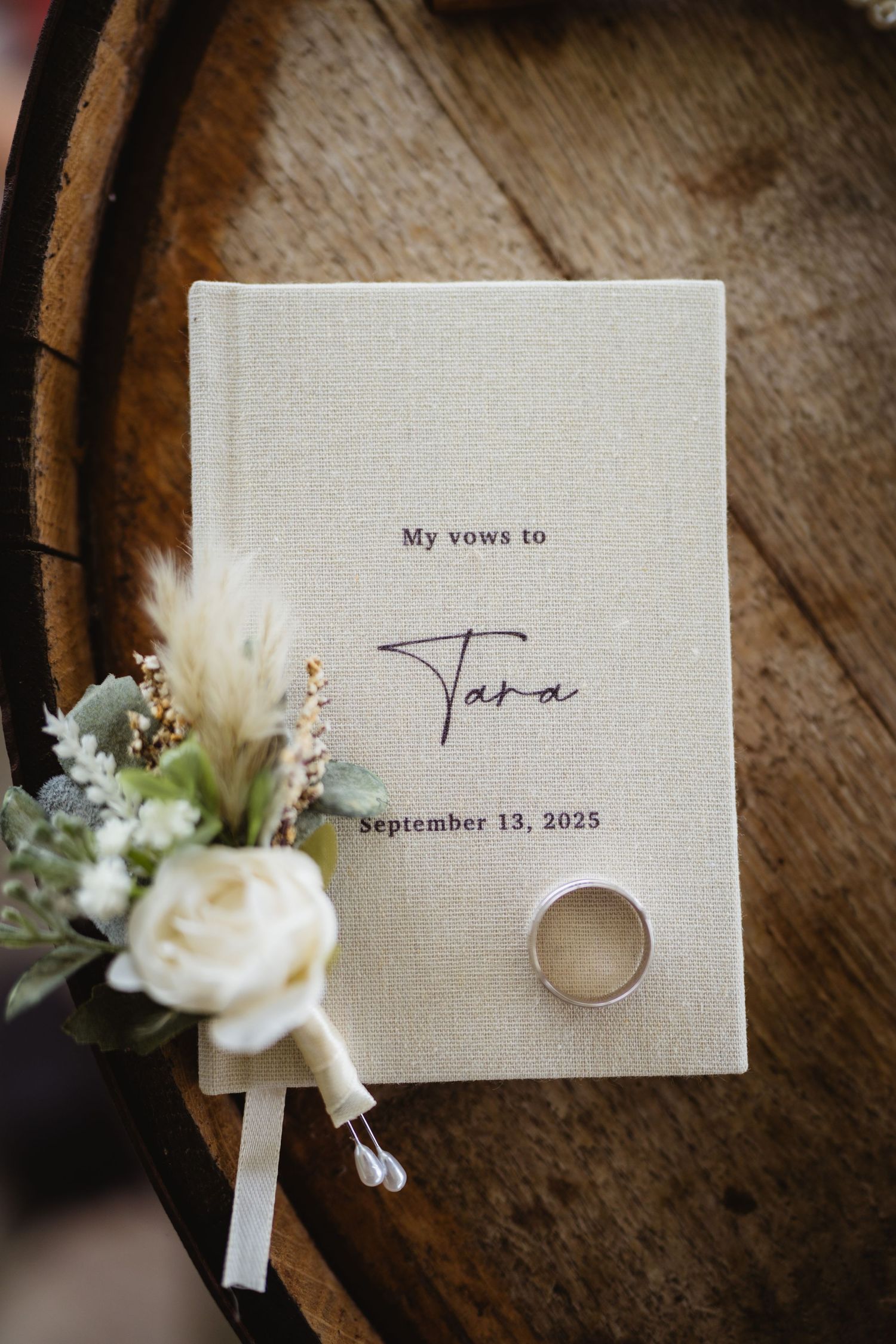 Wedding vows and floral boutonniere details arranged on rustic wooden surface show delicate white flowers and handwritten text.