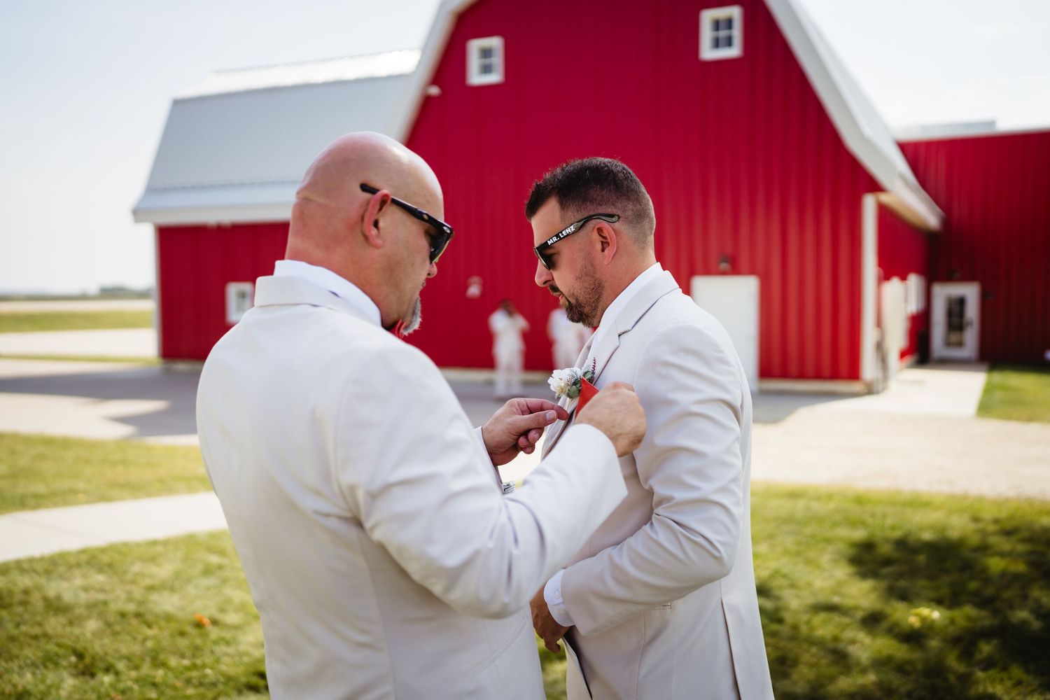 Two people in matching white shirts share a tender moment in front of a red barn on their wedding day.