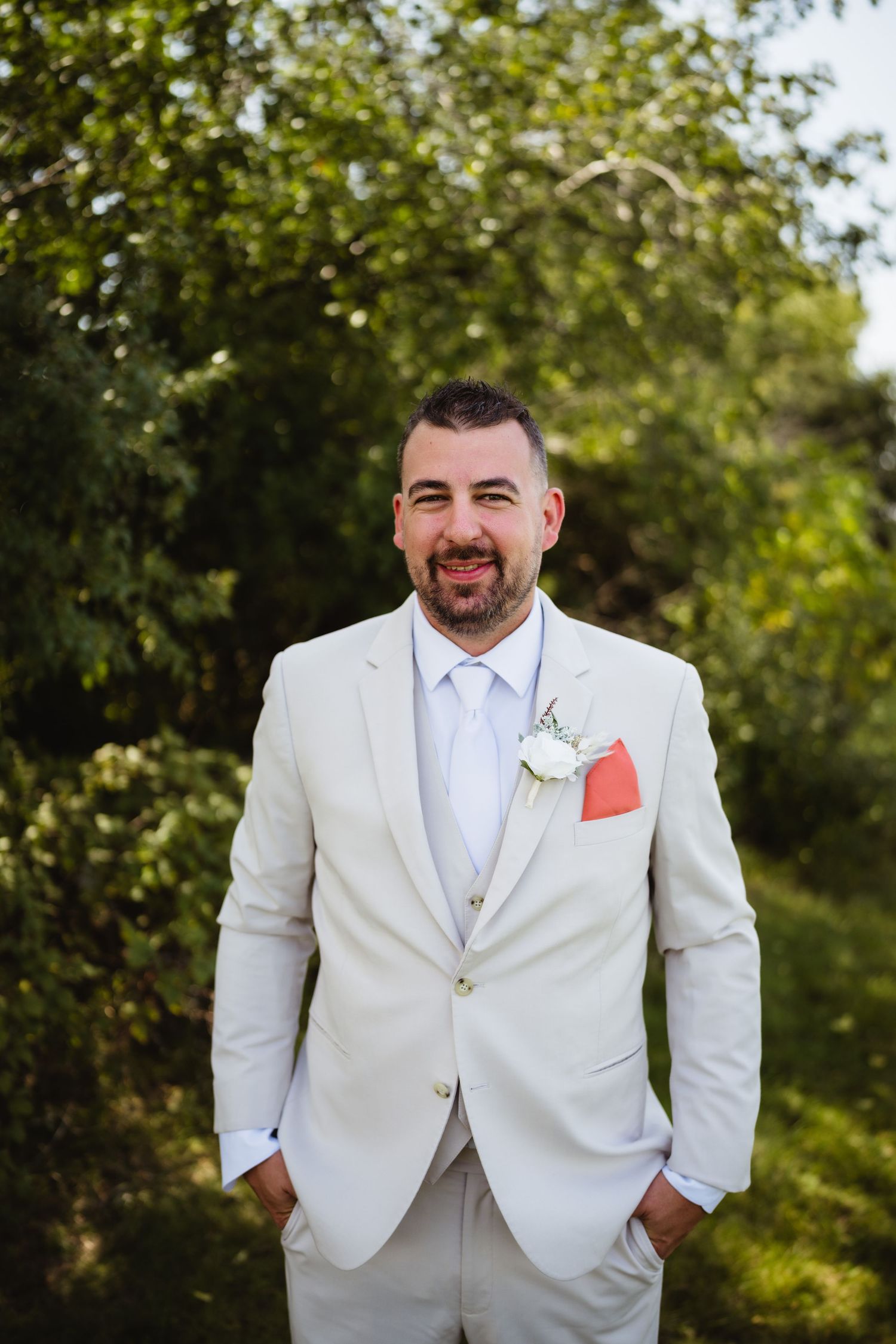 A person in a light beige suit with a coral pocket square poses outdoors against a green leafy background.