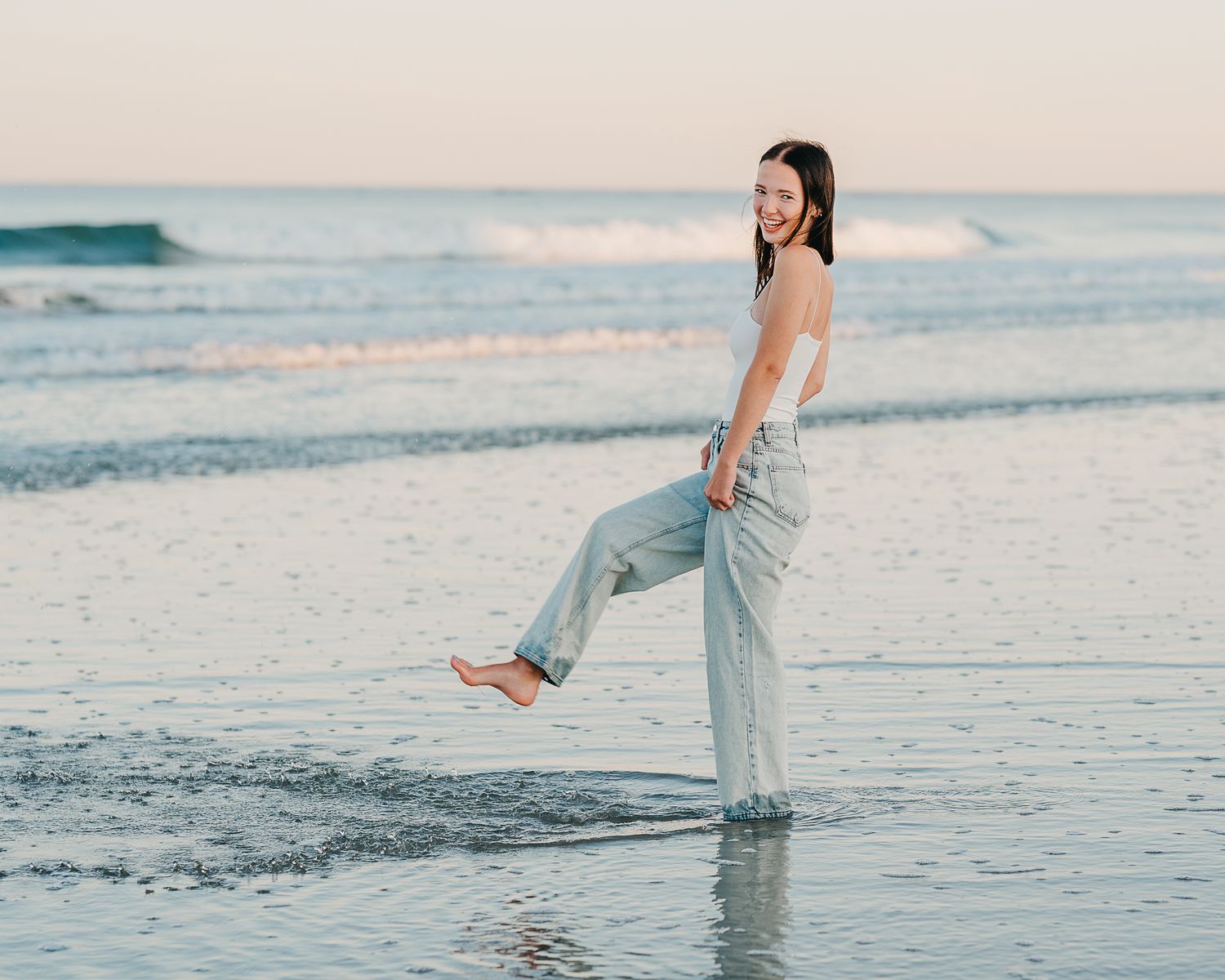 A person in casual attire poses joyfully on a serene beach during sunset with warm lighting and reflective sand.