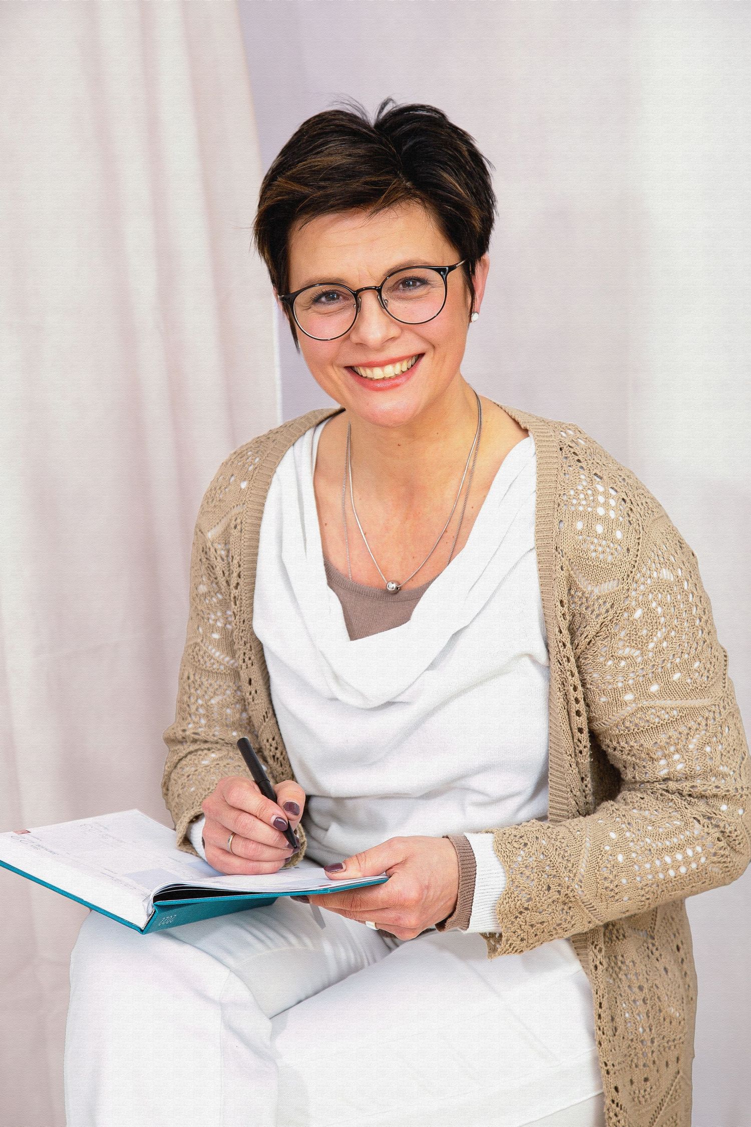Woman in beige cardigan and white top takes notes while sitting comfortably on couch.