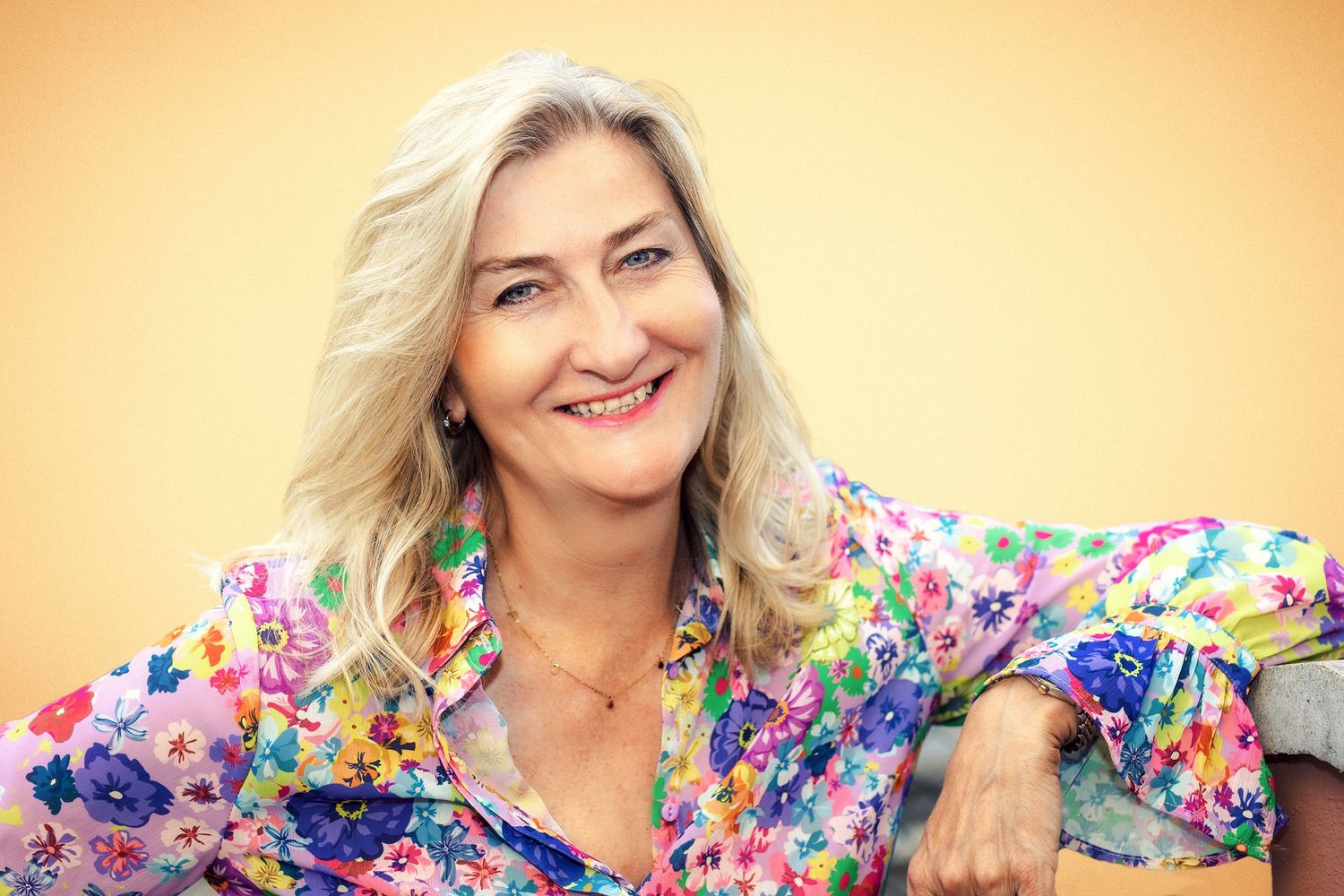 A cheerful person wearing a colorful floral blouse smiles warmly against an orange background.