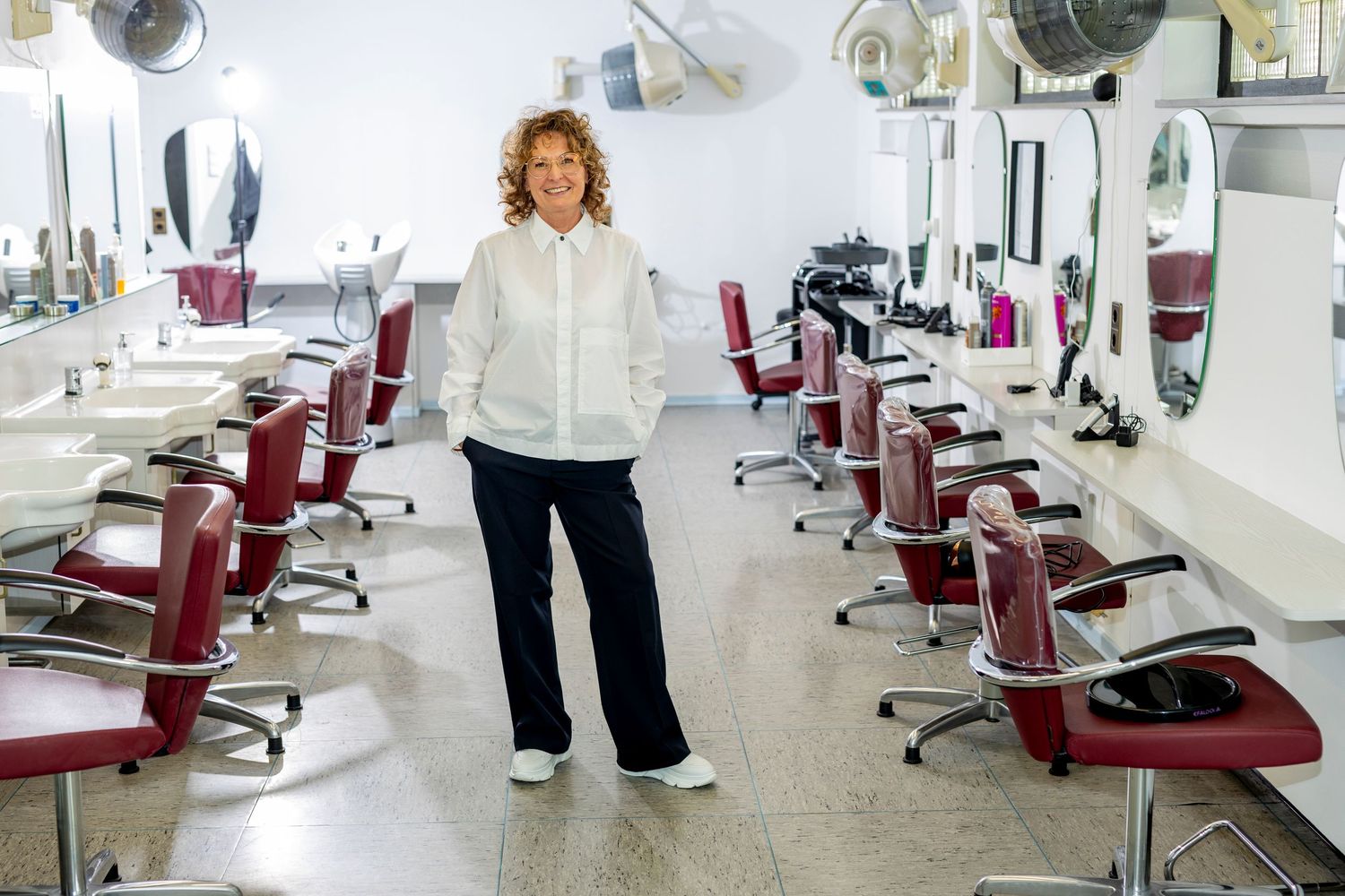 A person stands in an empty salon surrounded by red chairs and styling stations with mirrors.