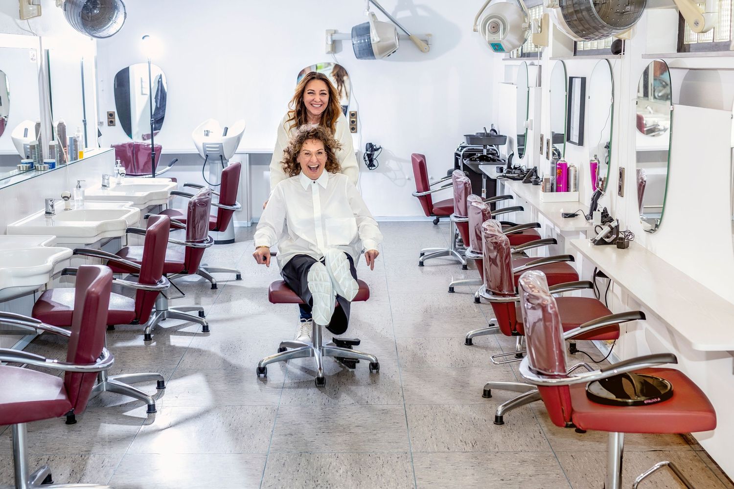 A modern hair salon interior shows empty styling stations with red chairs and mirrors in a bright white space.
