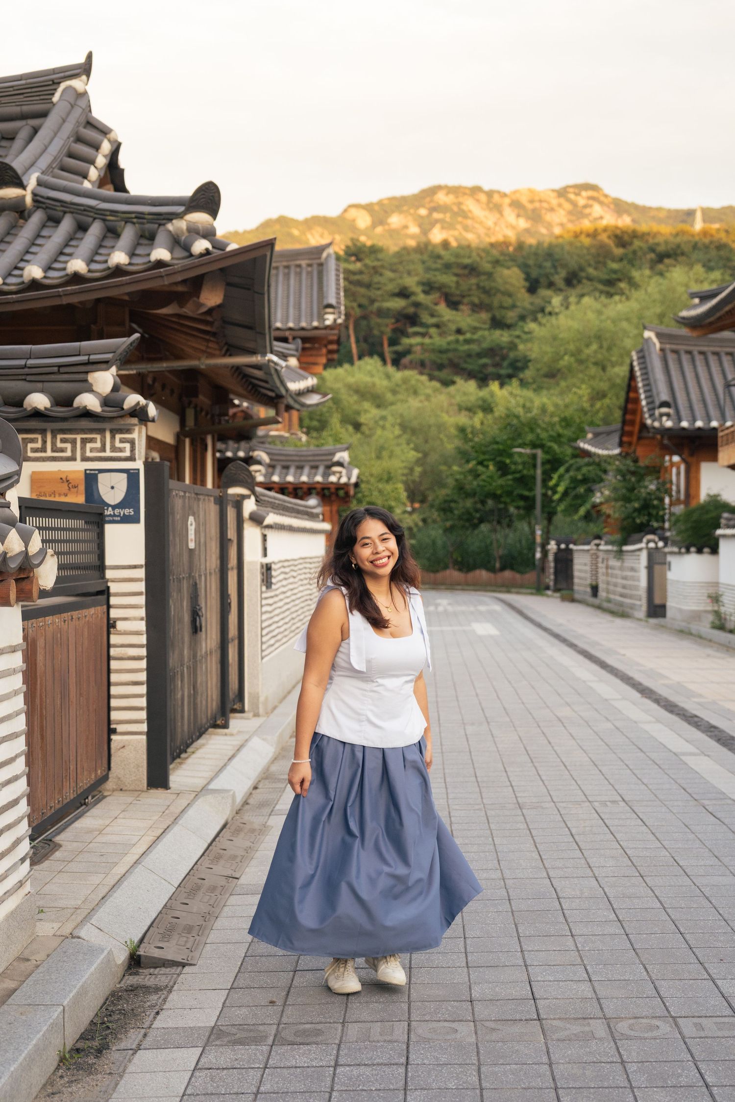 Person in white top and blue maxi skirt walking on stone path with traditional Korean architecture and mountains in background.