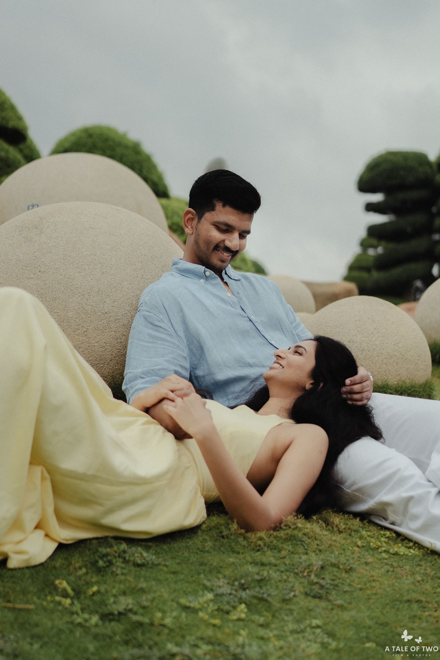 A romantic couple relaxes on grass near topiary gardens, with her in a yellow dress and him in light blue.