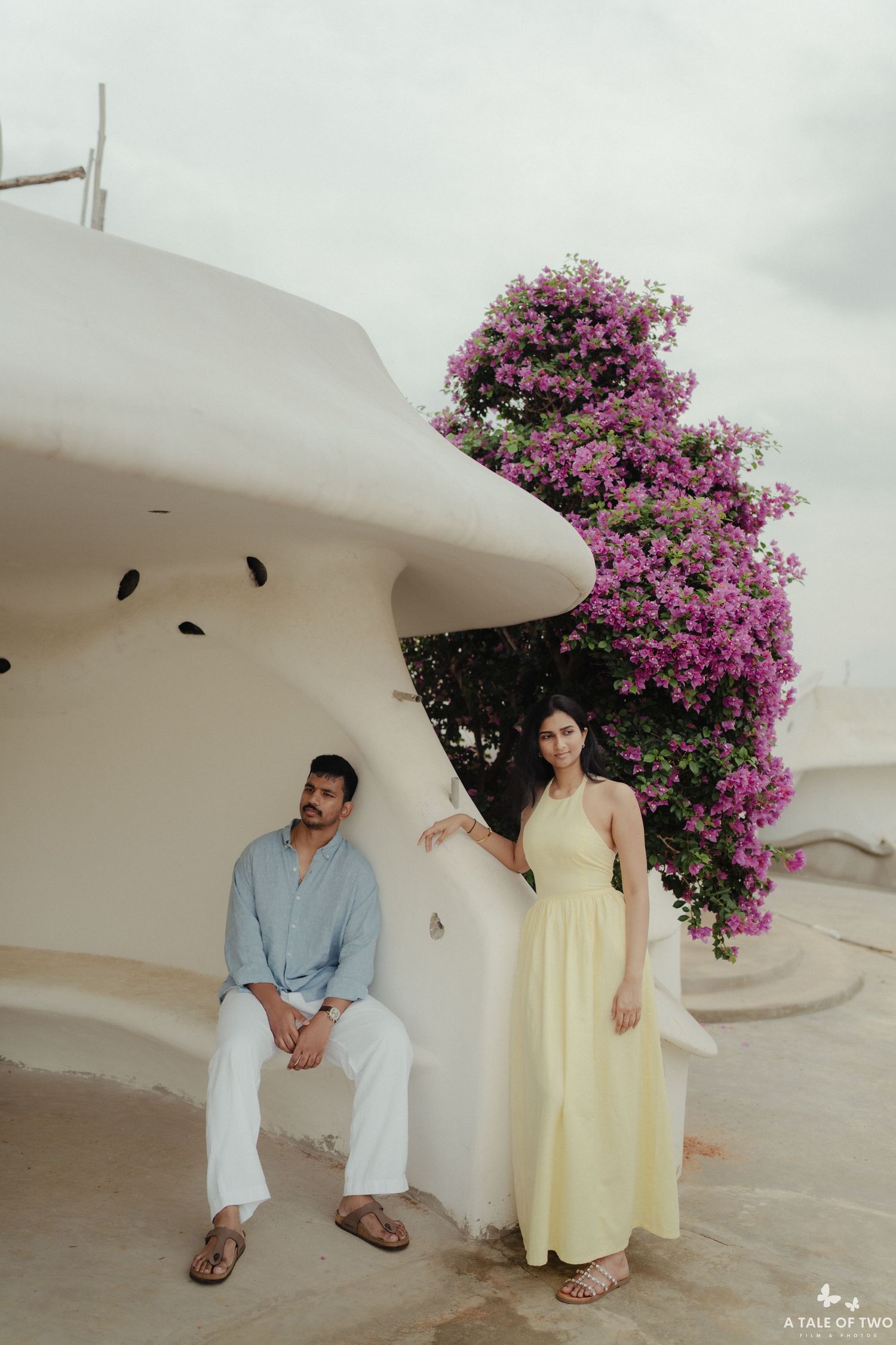 A romantic couple poses against white architecture with blooming pink bougainvillea flowers in a Mediterranean setting.