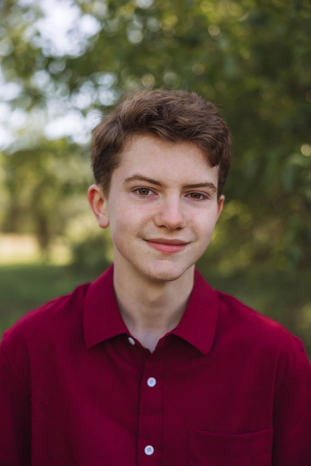 A person in a burgundy polo shirt poses against a soft-focus natural background.