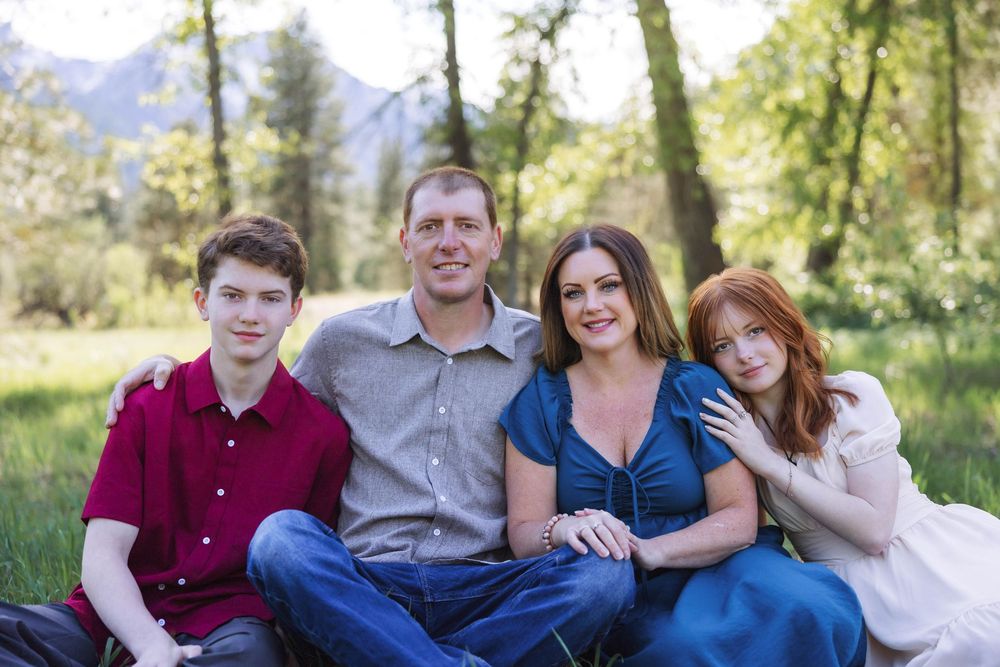 A family group sits together on grass with trees and mountains visible in the distance.