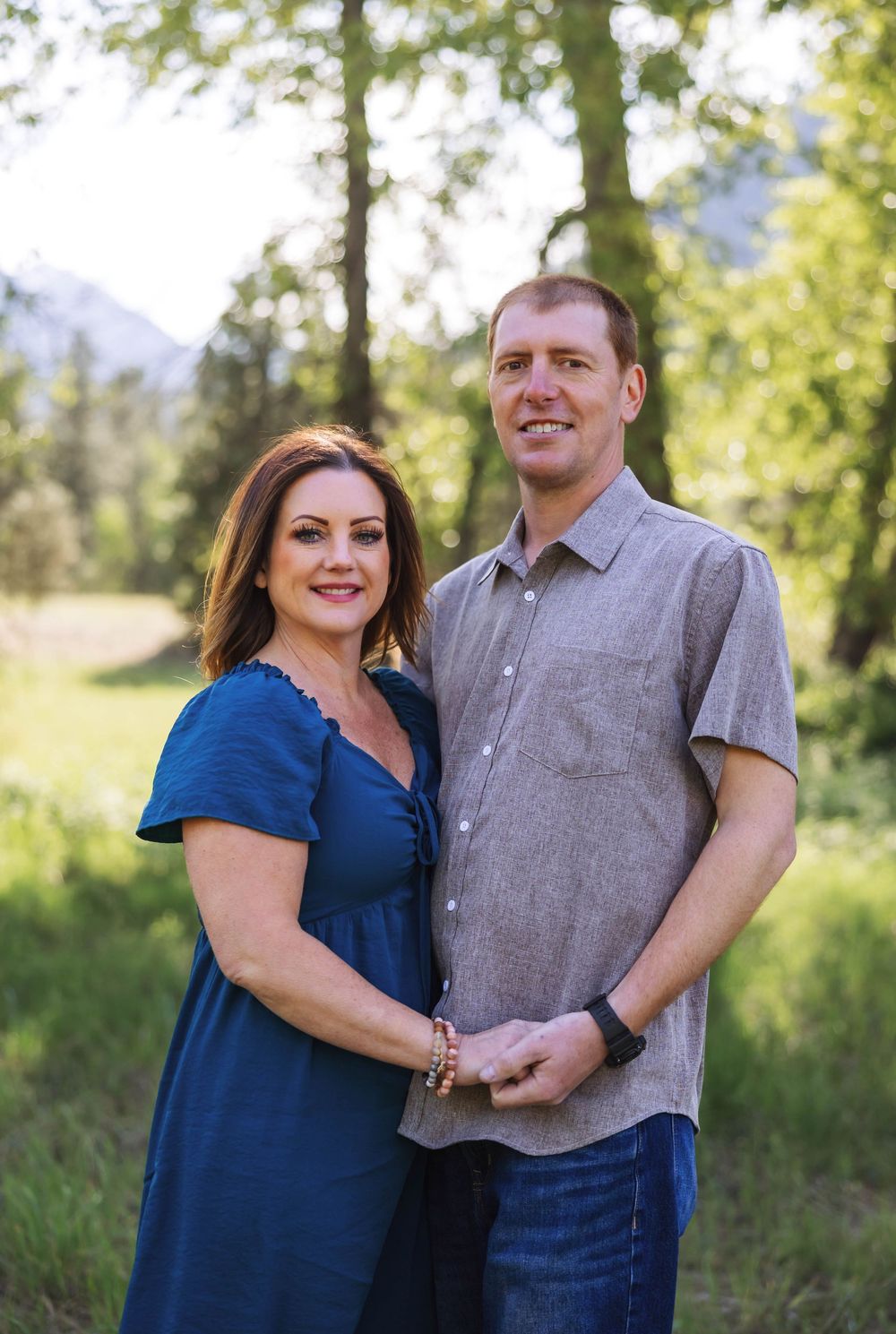 A couple in blue and gray outfits poses together outdoors in a natural setting with trees and mountains.