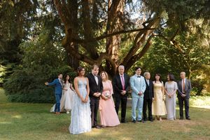 Wedding party gathered for group photos on grass under large trees in natural outdoor setting.