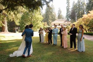 Wedding party gathered outdoors in a beautiful garden setting at golden hour with sunlit trees in background.