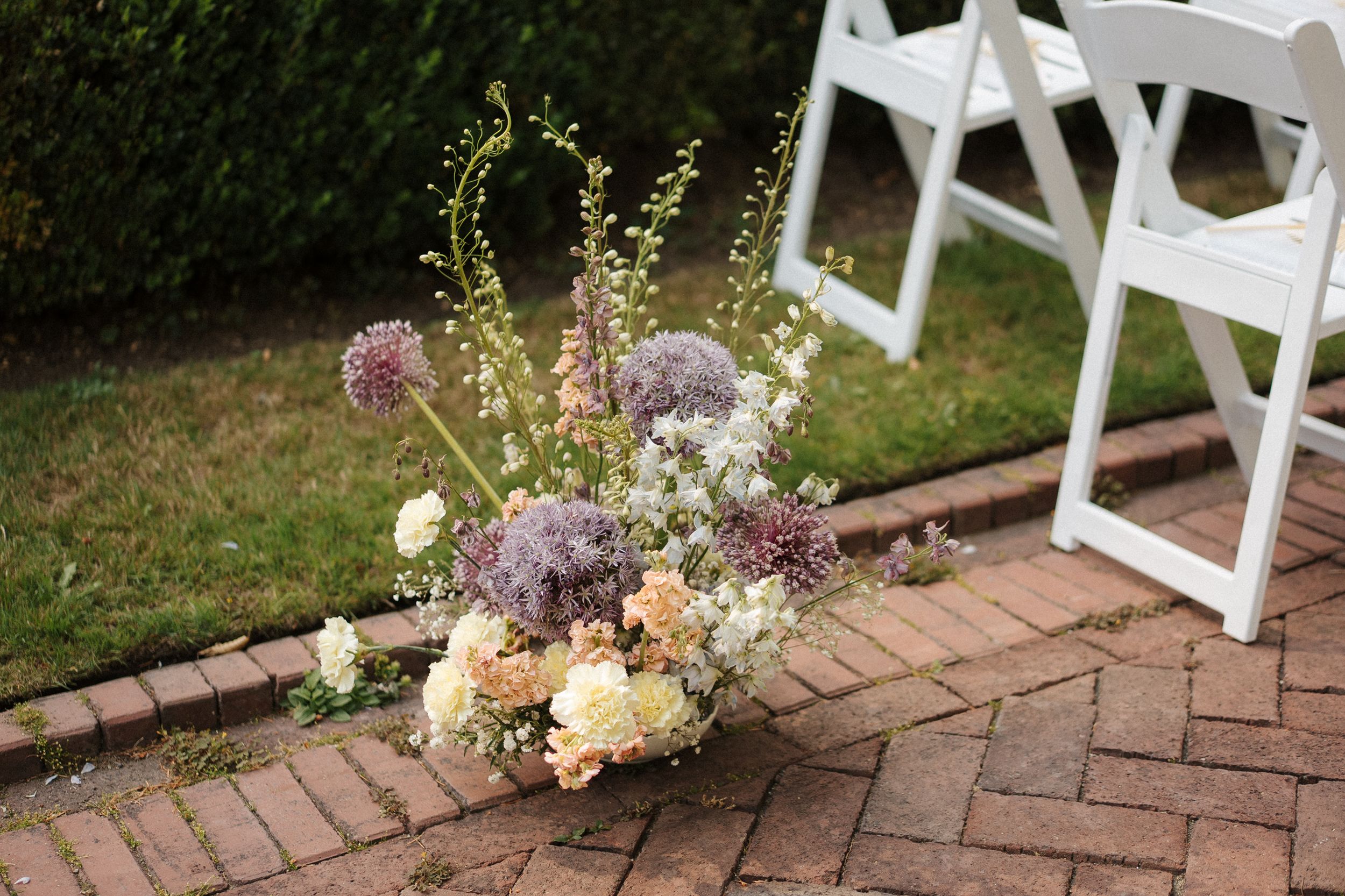 Elegant outdoor wedding venue with rows of white folding chairs set up on a brick patio bordered by flowers and grass.