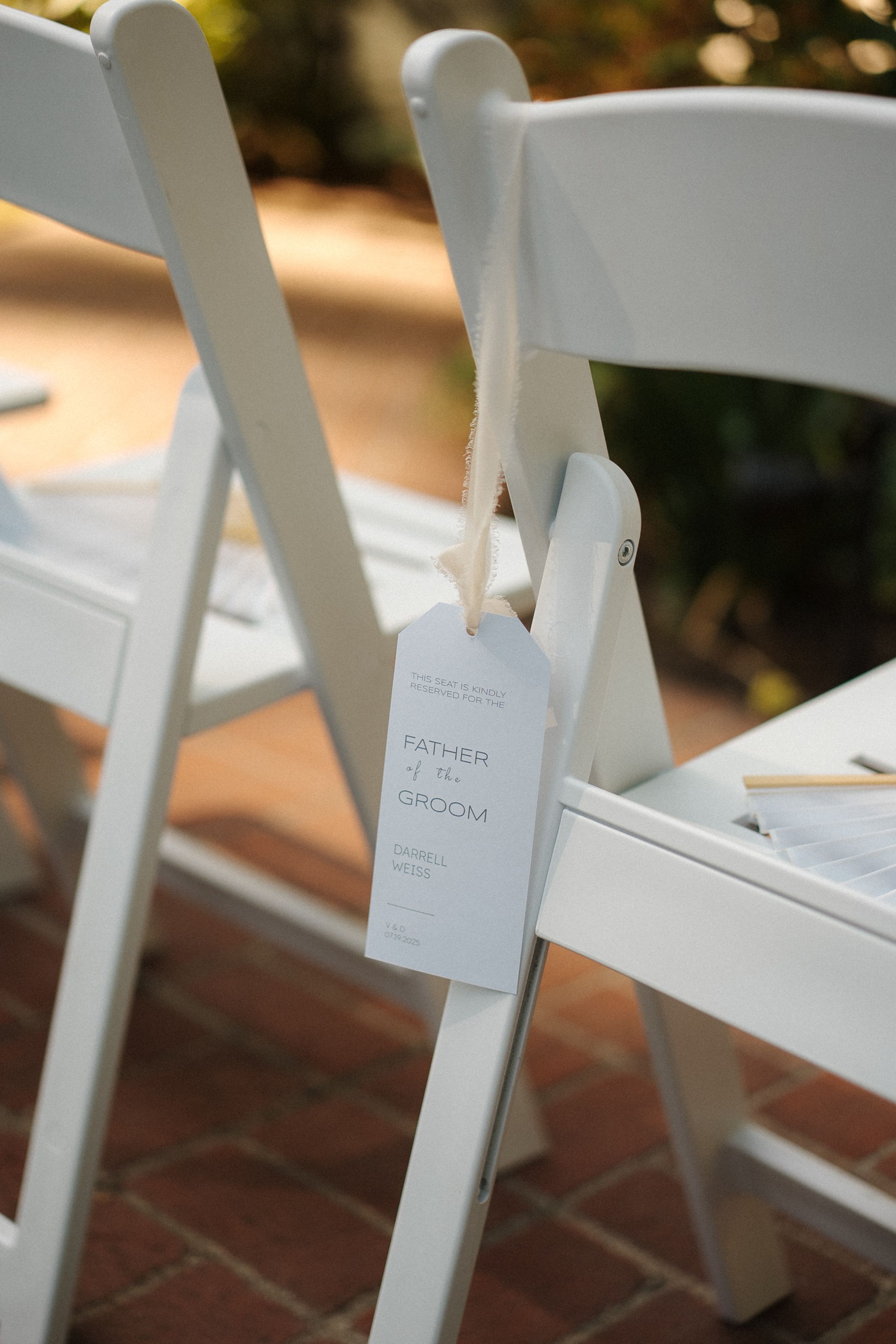 White folding chairs with small name cards attached line up at an outdoor wedding ceremony on brick pavement.