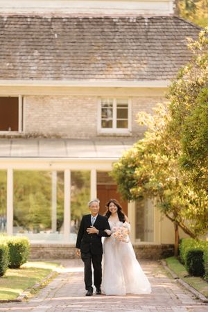 Two figures in wedding attire walk along a beautiful garden pathway at sunset with a white house in the background.