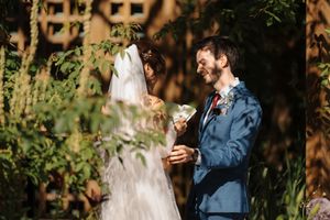 A bride in a white wedding dress and groom in a navy suit share an intimate moment in a rustic outdoor setting.