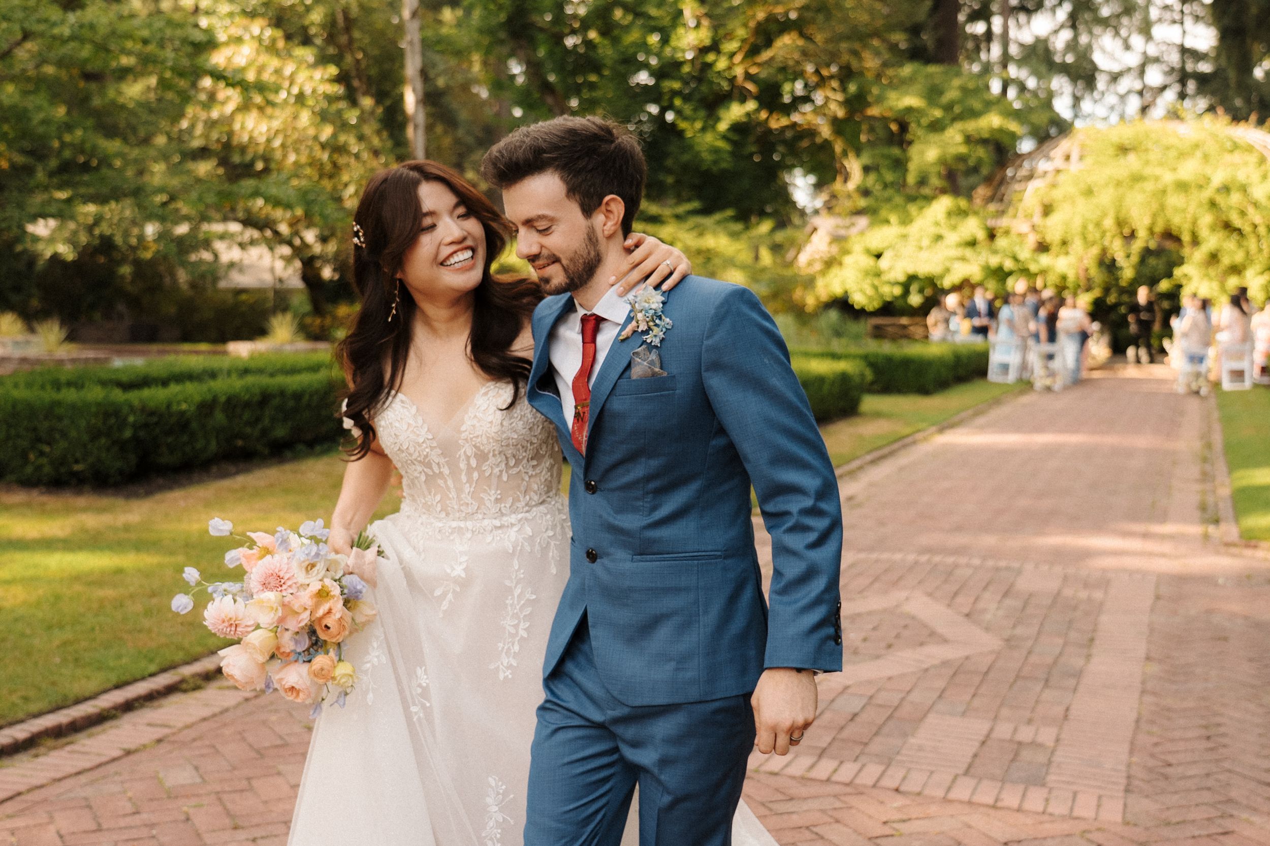 Happy newlywed couple walking and embracing on a brick pathway surrounded by lush greenery.