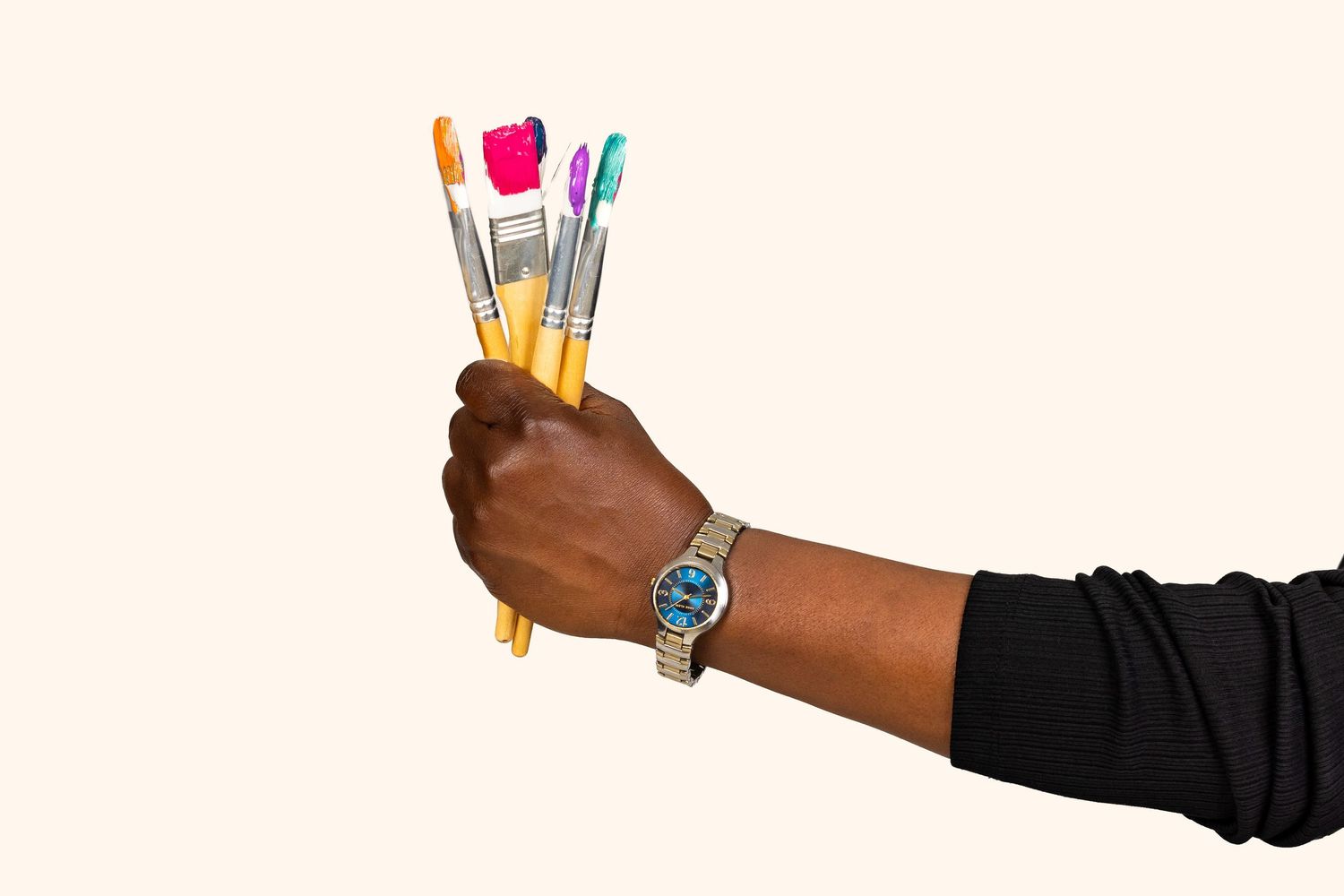Hand holding multiple colorful paintbrushes and pencils wearing a gold watch against white background.