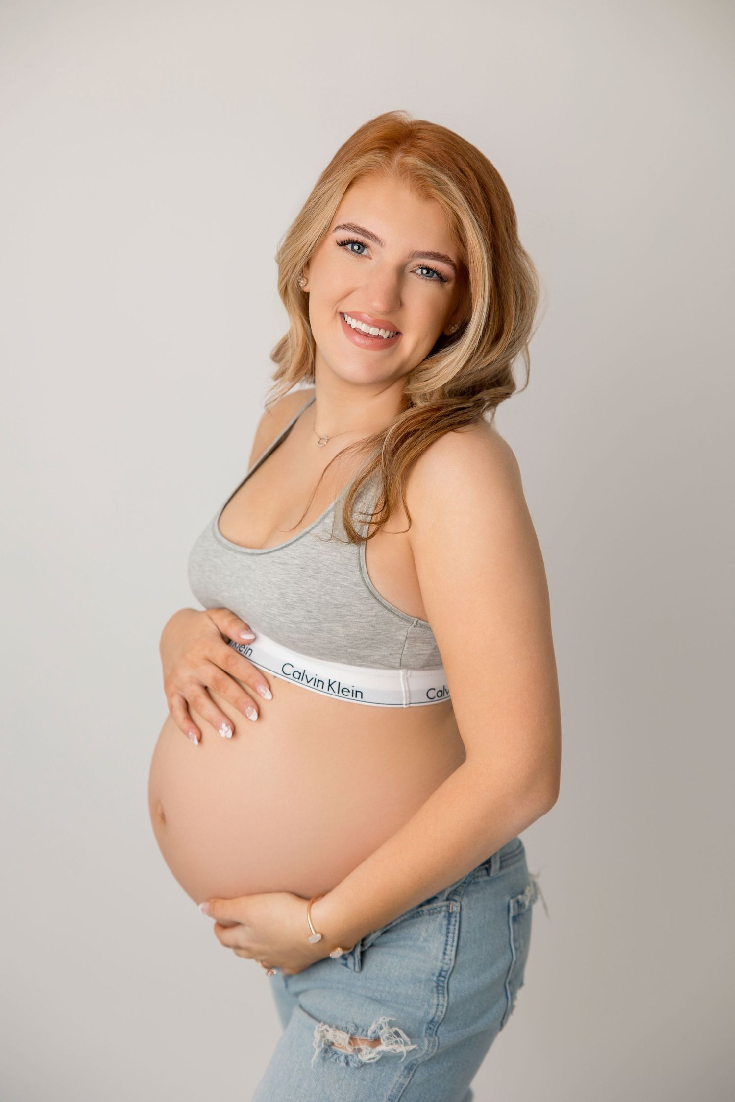 Maternity photoshoot showing a pregnant woman in a gray crop top and ripped jeans against a light background.