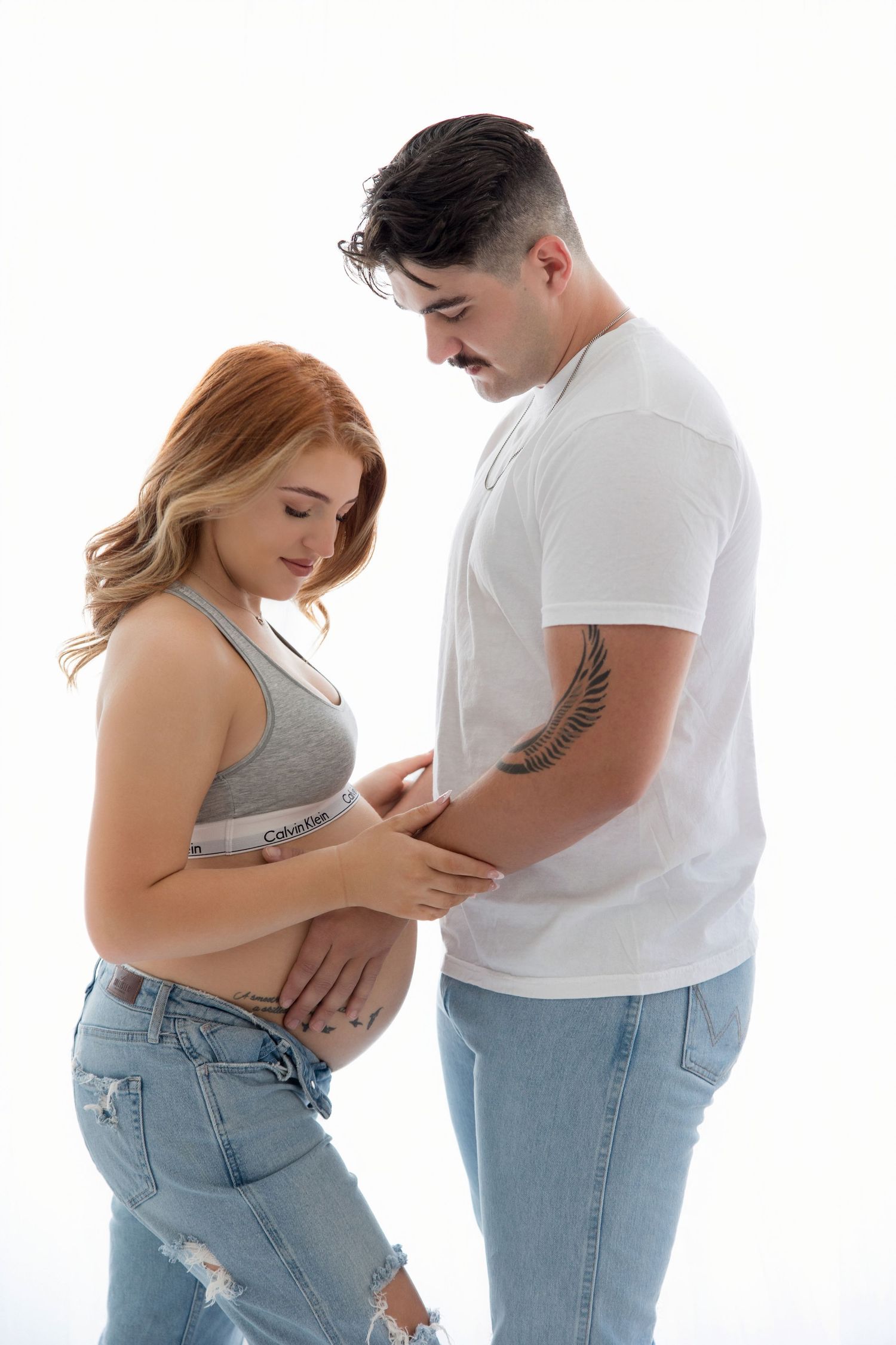 Couple embracing during pregnancy photoshoot wearing casual attire against white backdrop.
