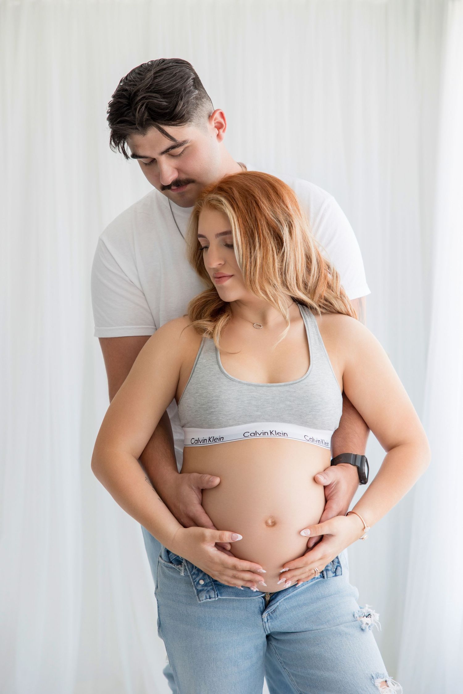 Intimate maternity photo series showing loving embrace of expecting couple in casual attire against white backdrop.