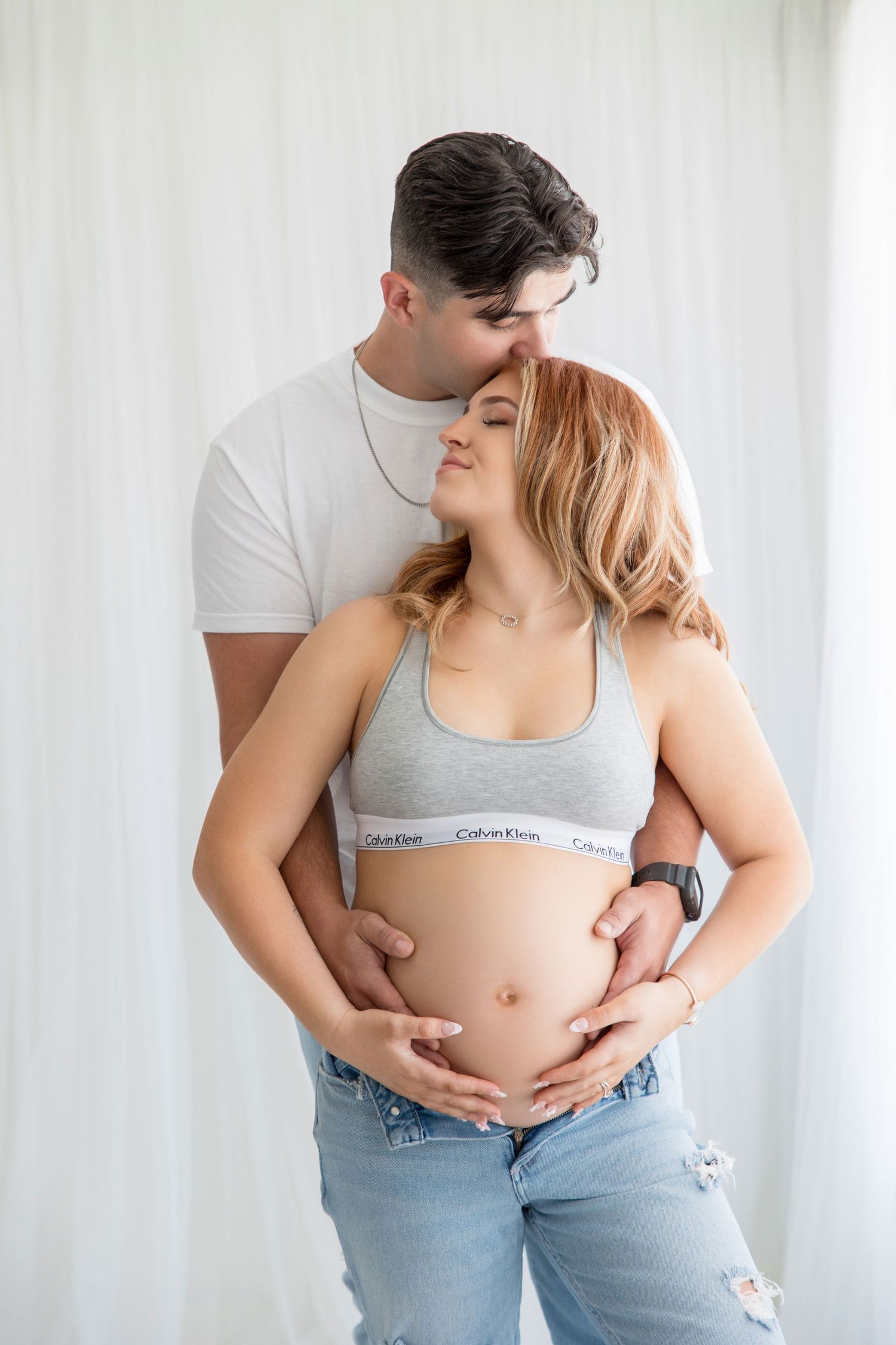 Intimate maternity photos of couple in white and gray casual wear against light background.