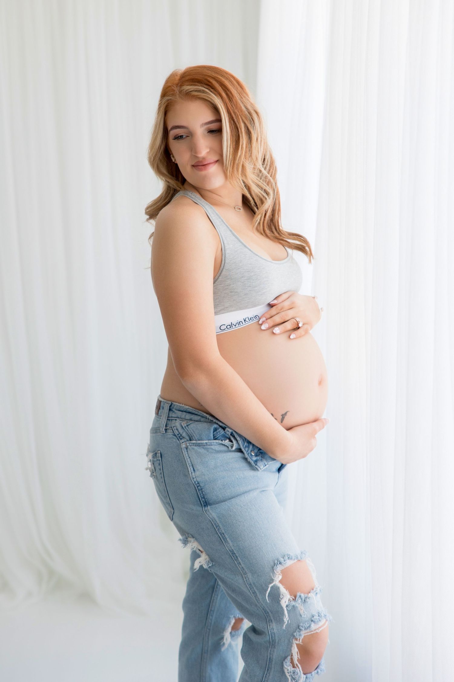 A maternity photo with casual style showing ripped jeans and a white top against a light curtained backdrop.