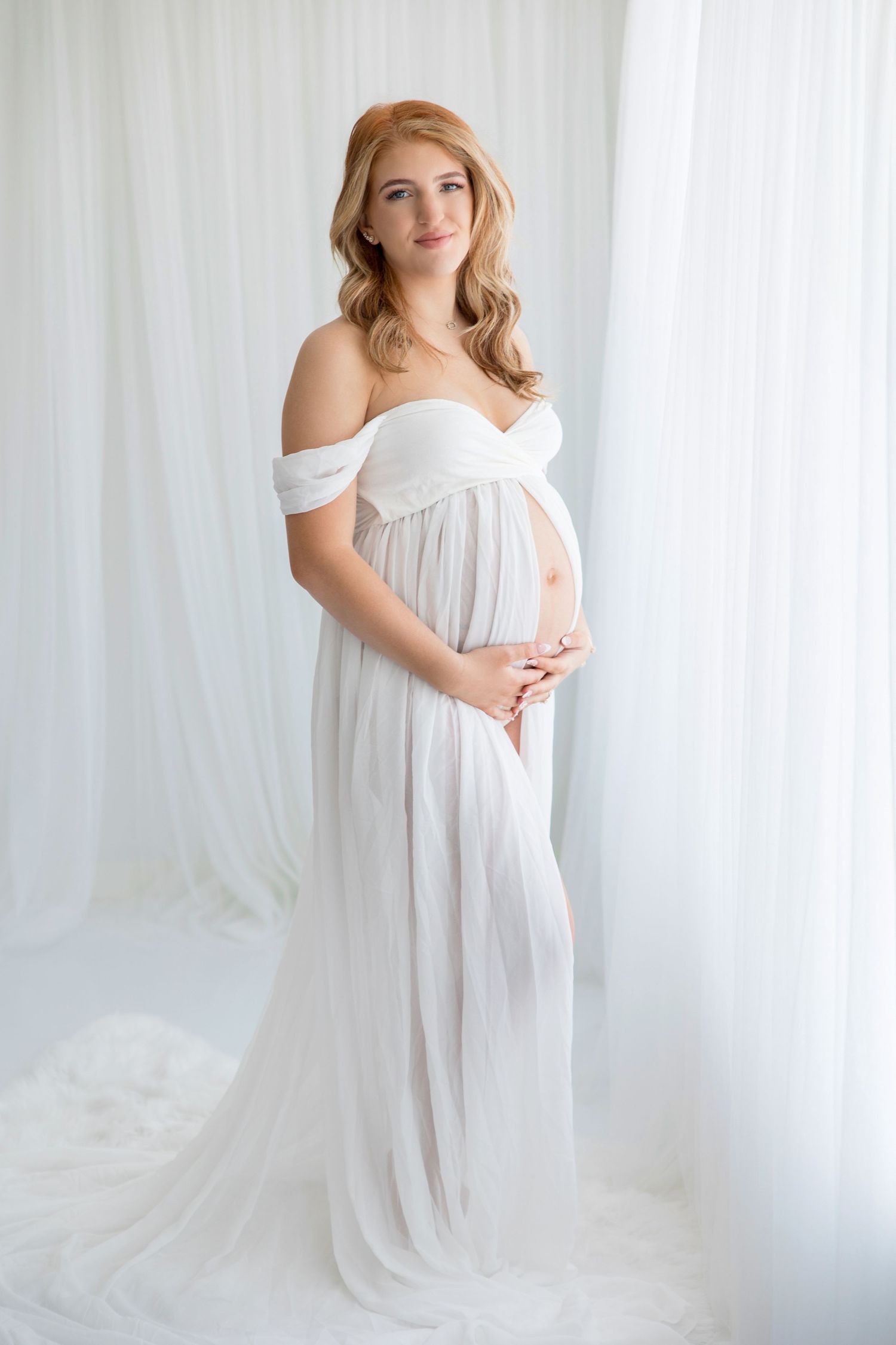 Romantic maternity portrait series with subject wearing delicate white off-shoulder dress against white backdrop.