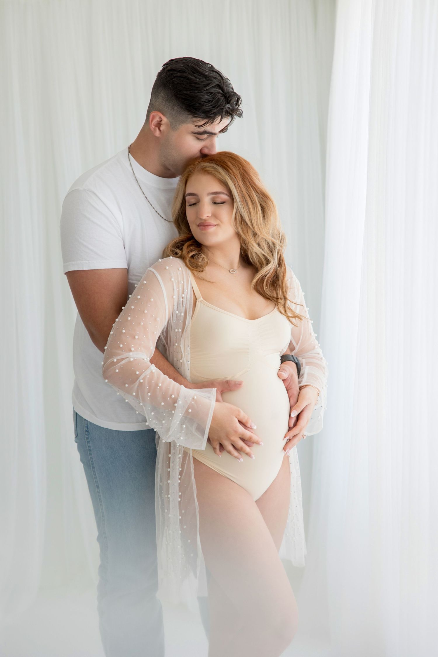 Intimate maternity photo session showing tender couple moments in soft white light and flowing lace.